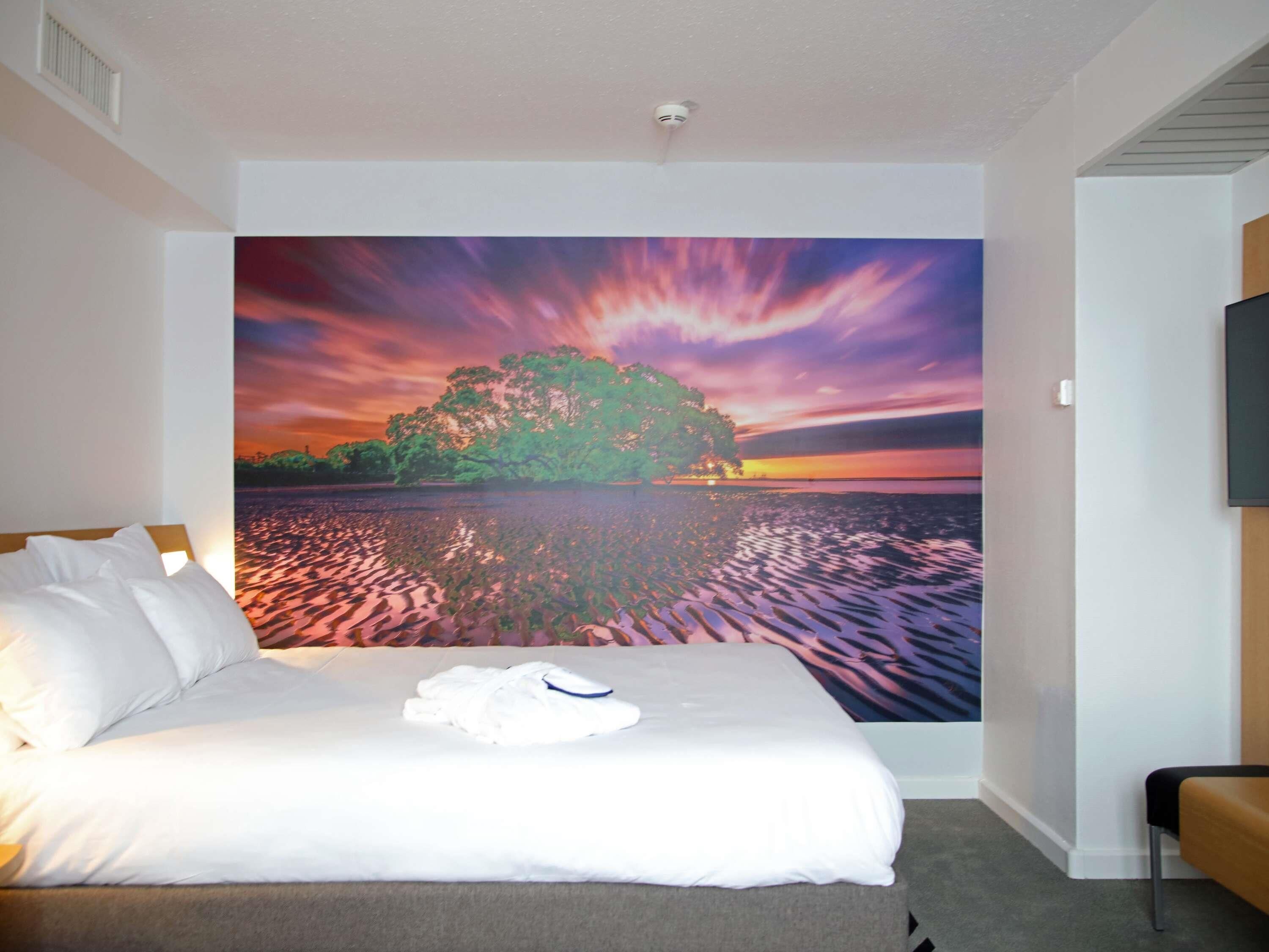 A hotel room with a bed, a flat-screen TV, and a large wall mural of a sunset over a lake.