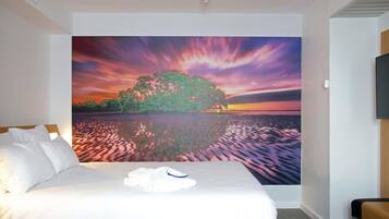 A hotel room with a bed, a flat-screen TV, and a large wall mural of a sunset over a lake.