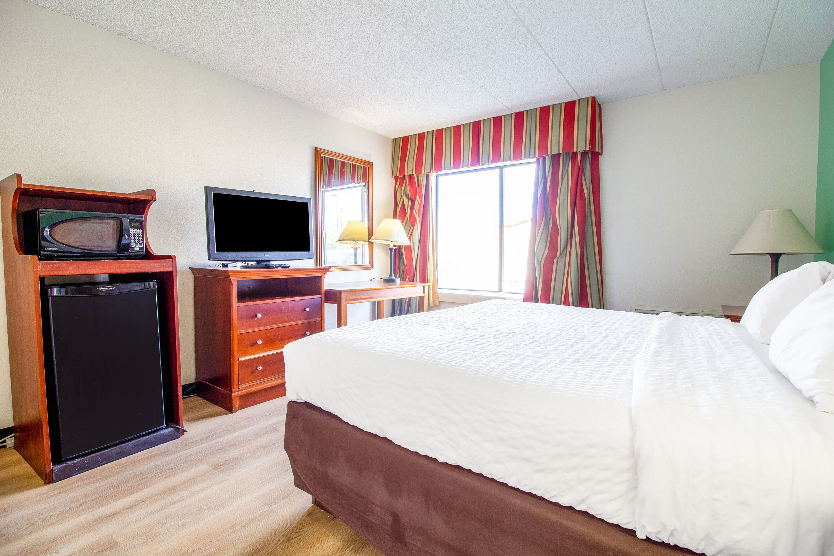 Una habitación de hotel con cama, televisión, refrigerador pequeño, escritorio con lámpara y ventana con cortinas.