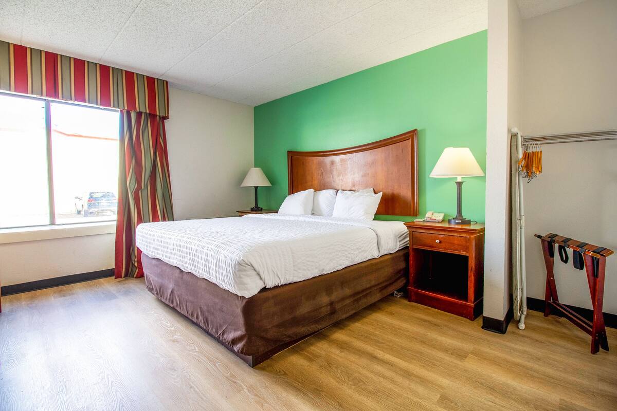 A hotel room with a large bed, a wooden nightstand, a lamp, and a window with curtains.