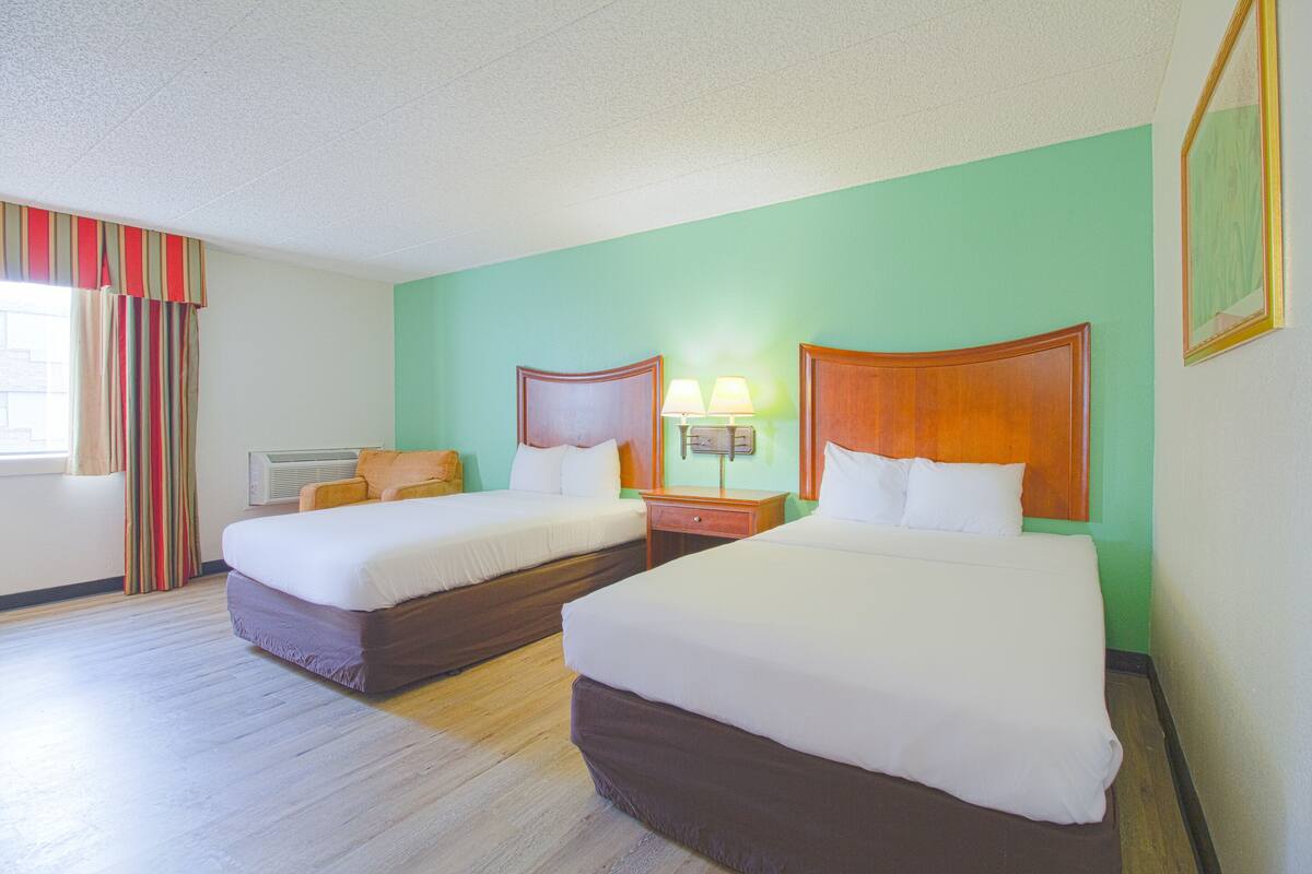 A hotel room with two beds, a nightstand with a lamp, a window with curtains, and a wall-mounted picture frame.