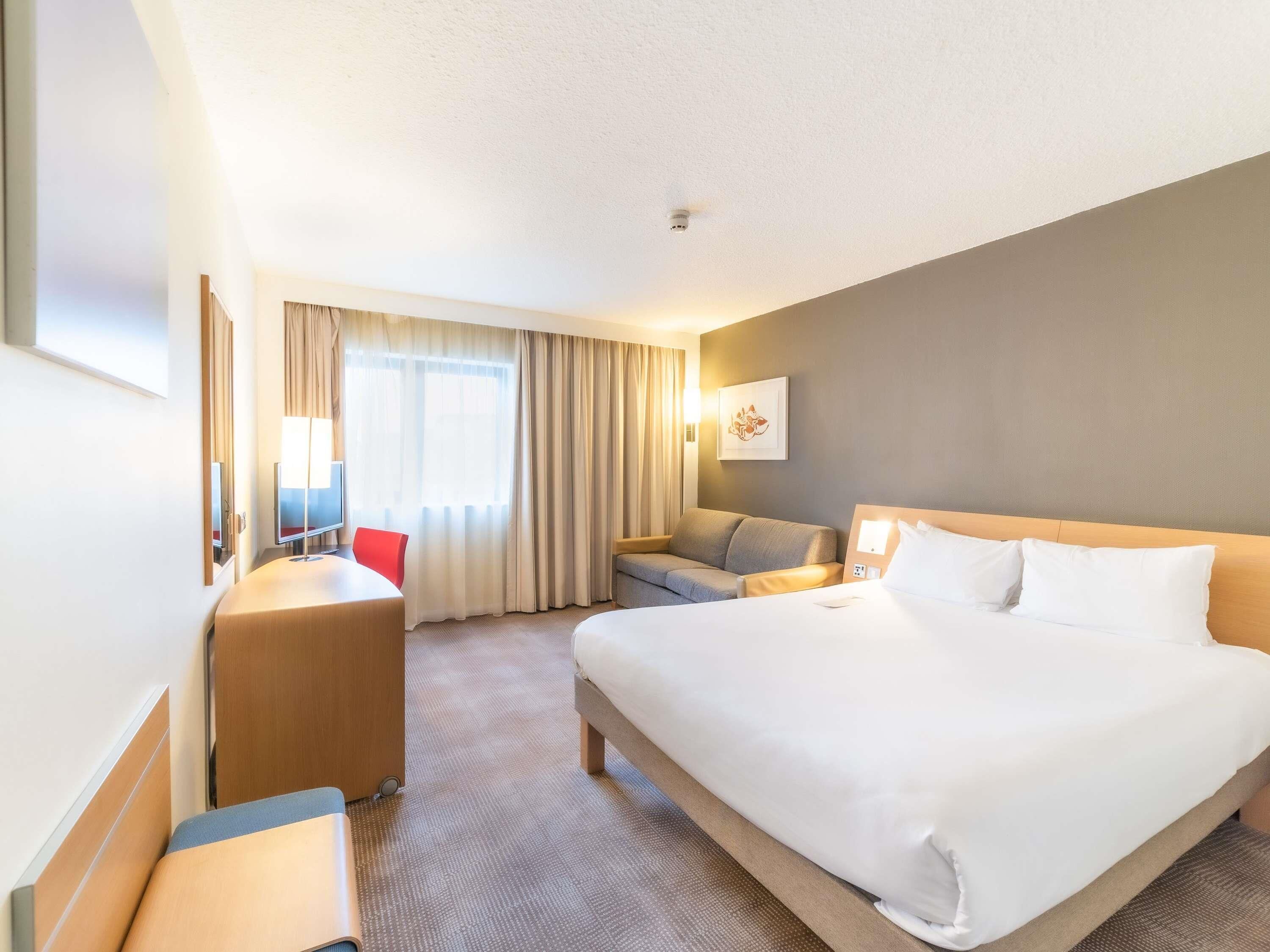 A hotel room with a large bed, a desk, a chair, a sofa, and a TV.