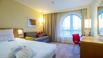 A hotel room with a large bed, a sofa, a desk, and a chair. There is a window with curtains and a view of the outside.