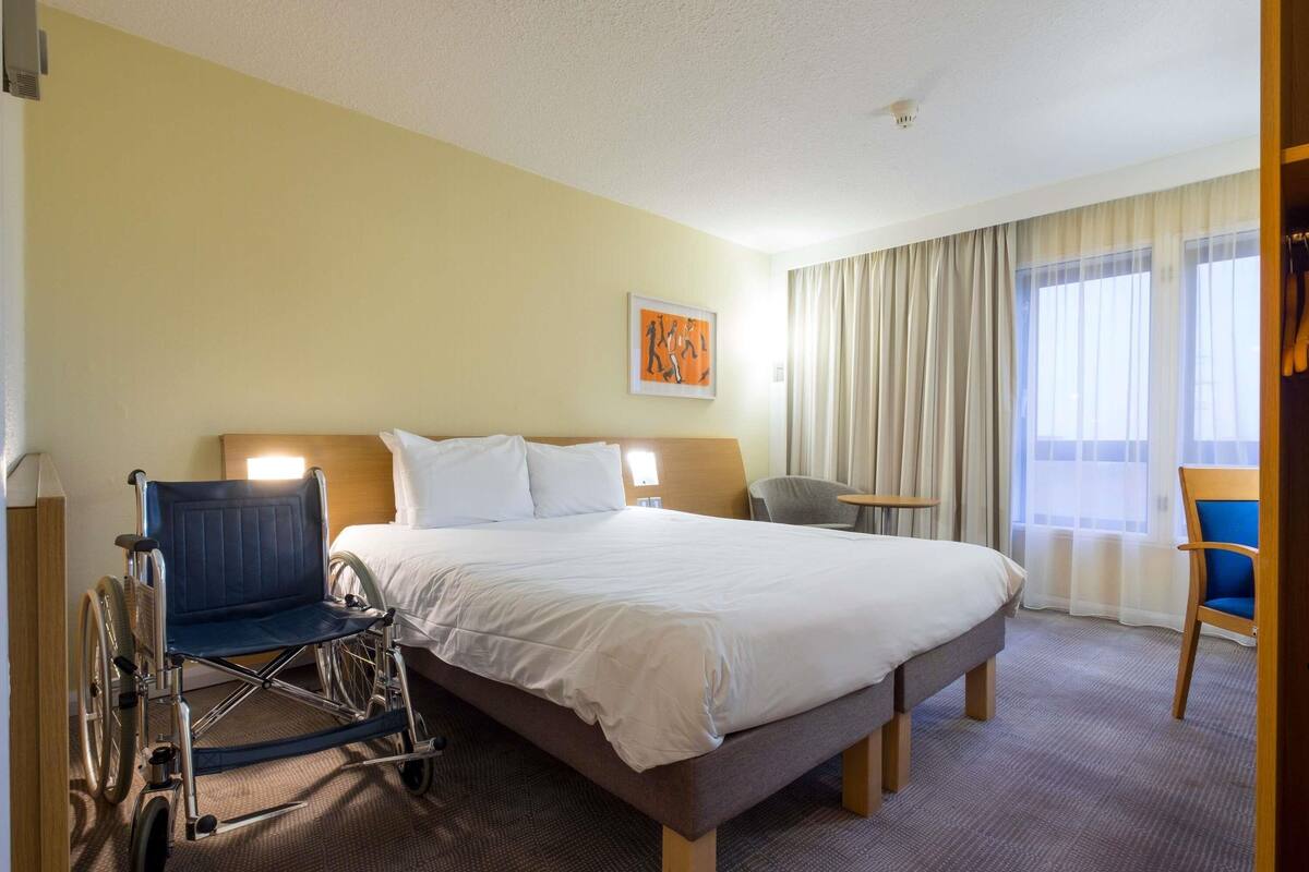 Een hotelkamer met een groot bed, een rolstoel, een klein tafeltje en een stoel.