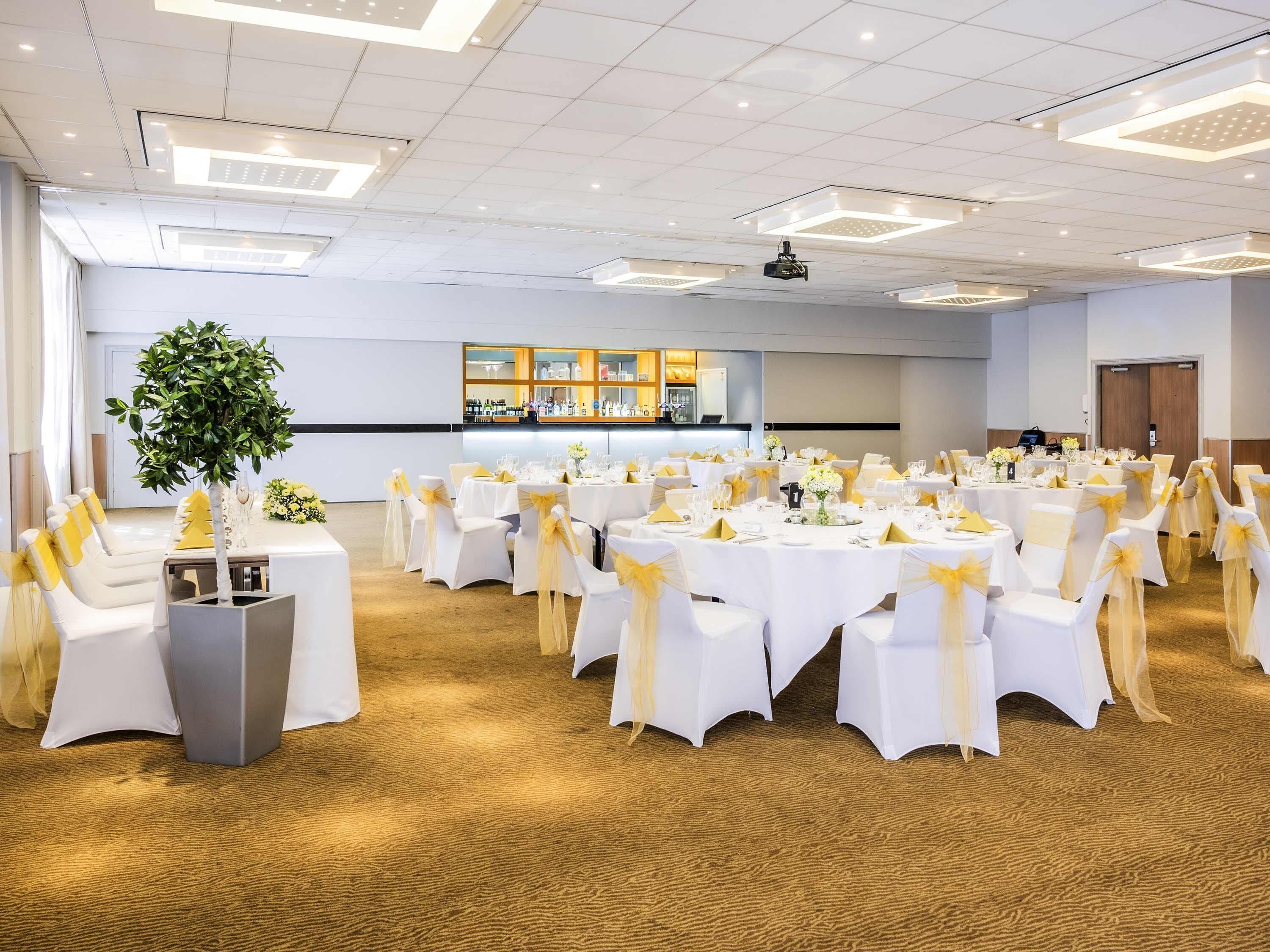 A banquet hall with round tables, white chairs with yellow sashes, a bar area, and a large plant.
