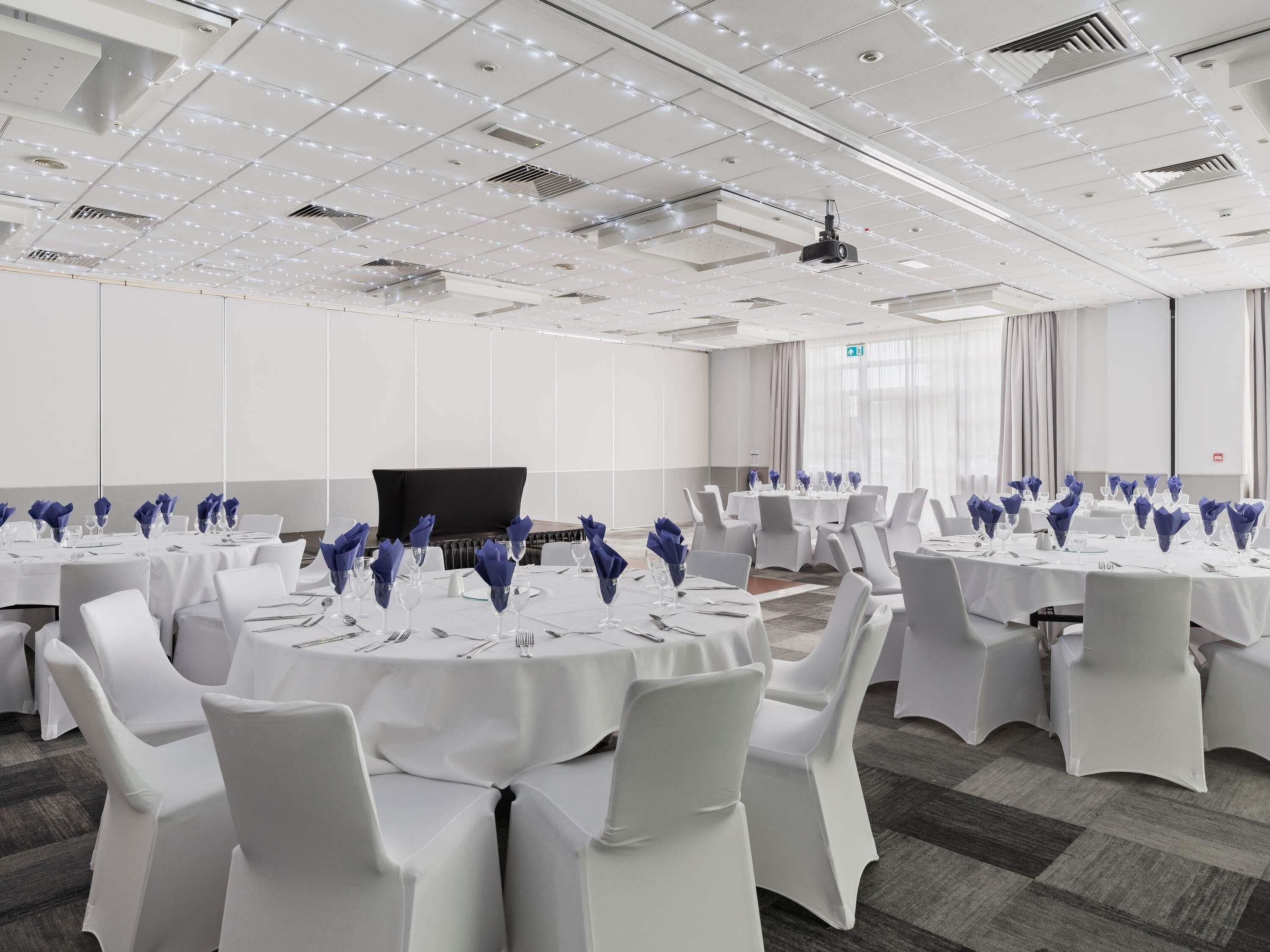 Un salón de conferencias con mesas dispuestas para una reunión, sillas blancas y un proyector en el techo.