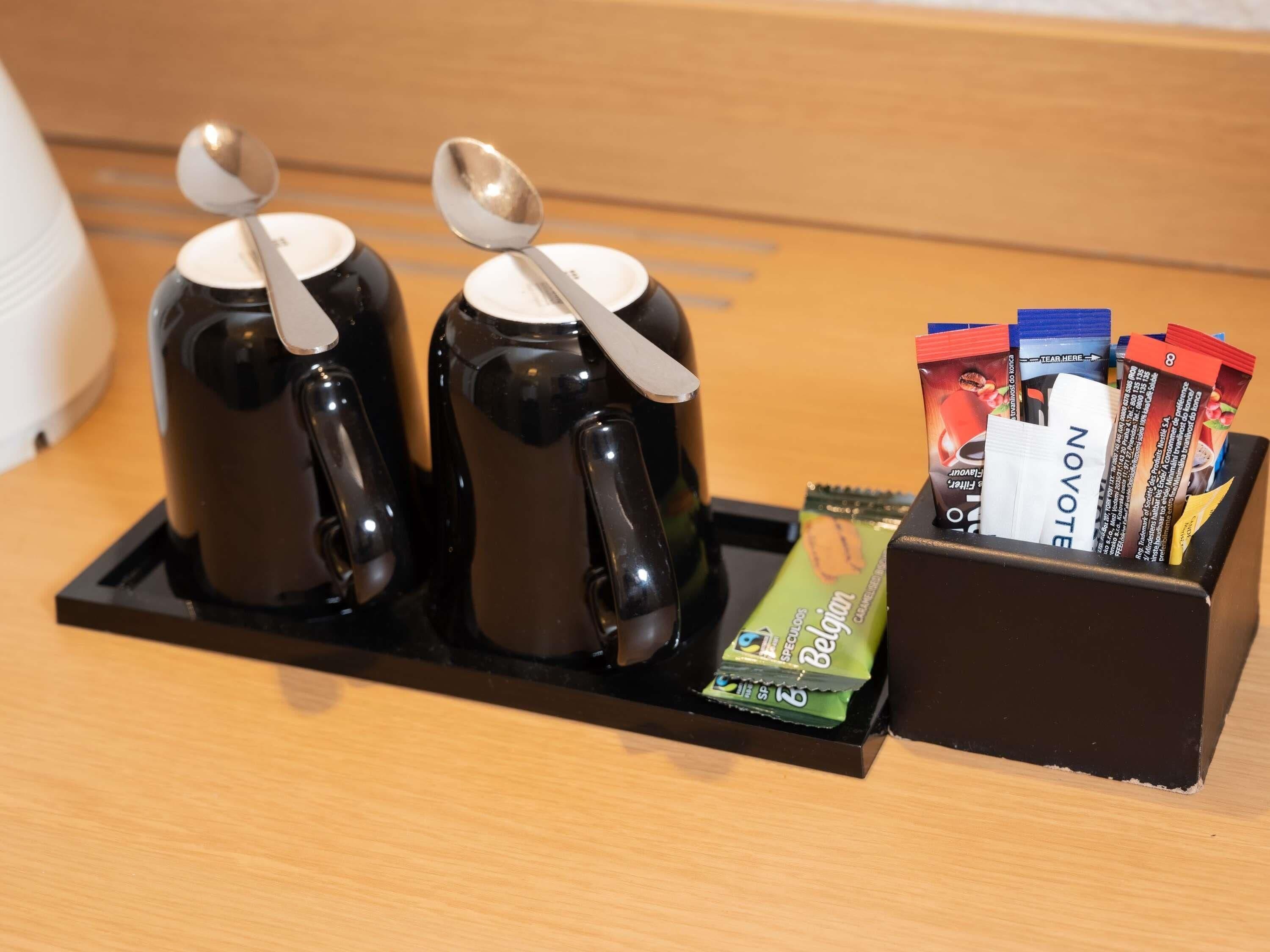 A tray with two black mugs, each with a spoon, and a box containing various snack packets.