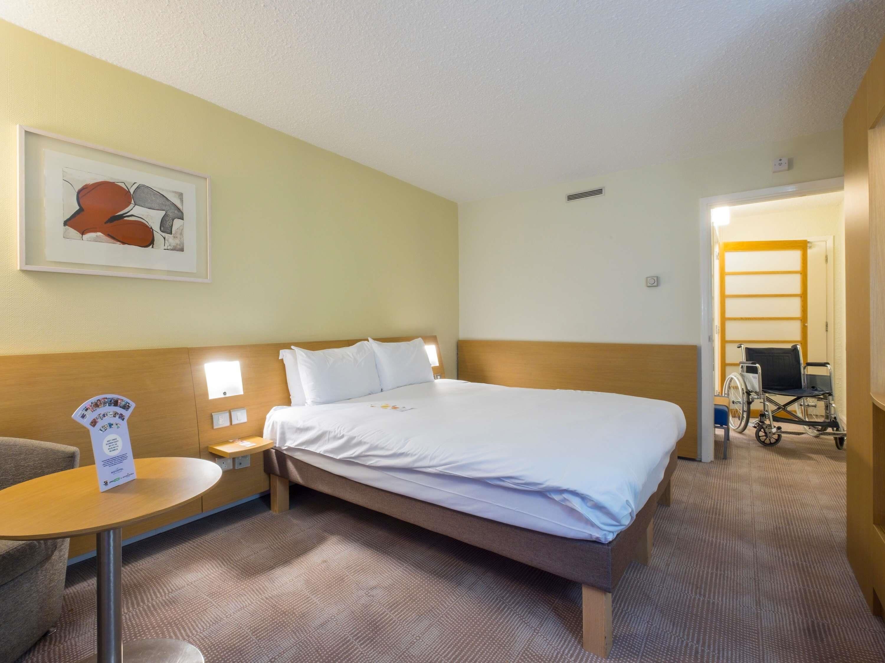 A hotel room with a bed, bedside tables, a chair, a small table, a framed picture on the wall, and a wheelchair in the hallway.