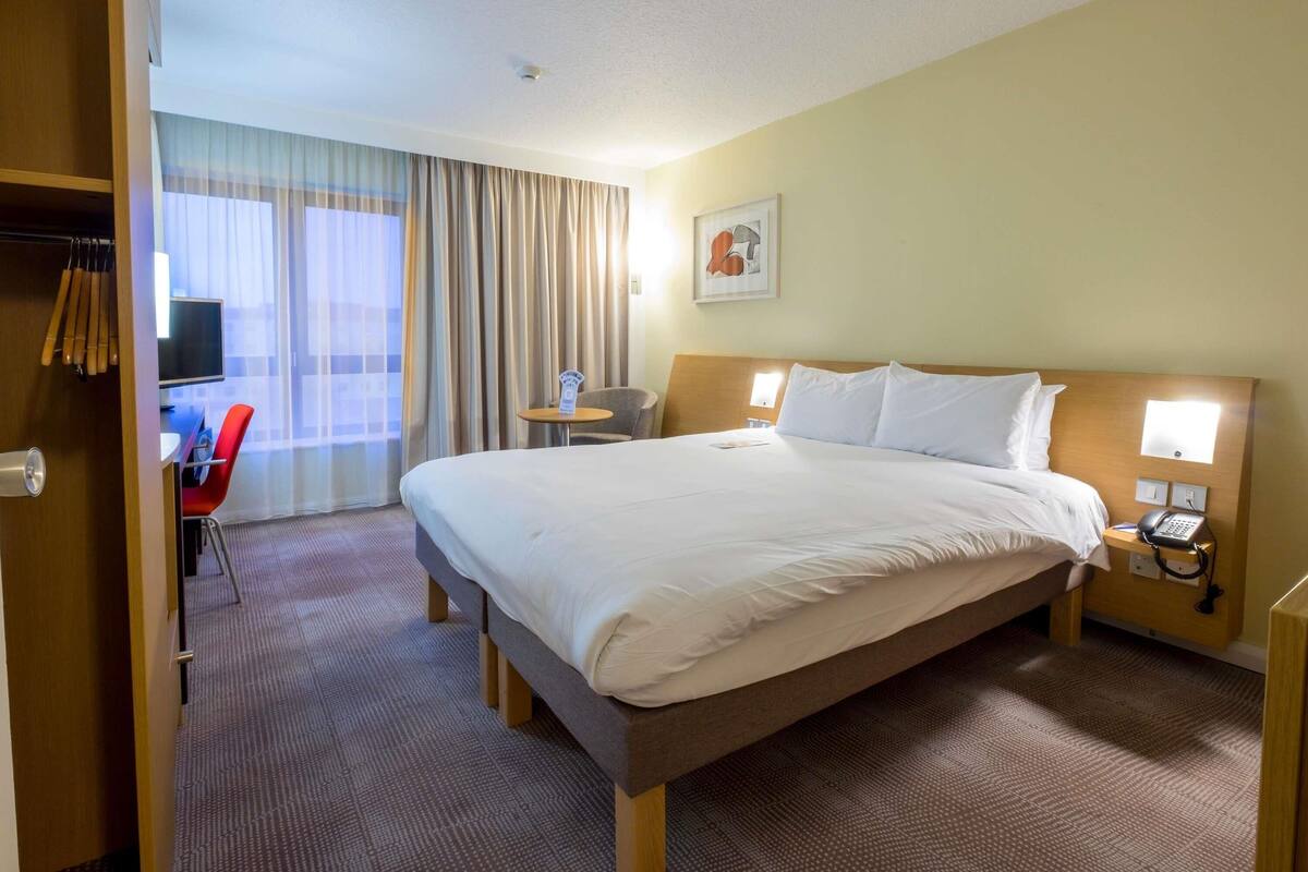 Een hotelkamer met een bed, nachtkastje, stoel, bureau, televisie en gordijnen.