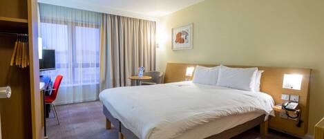 Une chambre d’hôtel comprenant un lit, une table de chevet, une chaise, un bureau, une télévision et des rideaux.