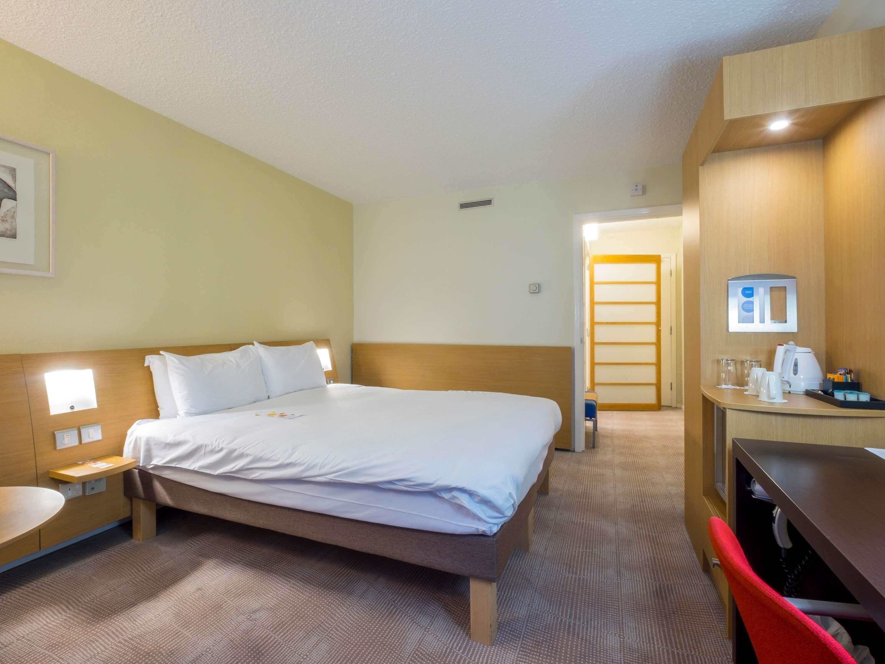 A hotel room with a bed, bedside tables, a desk with a chair, a mirror, and a door leading to another room.