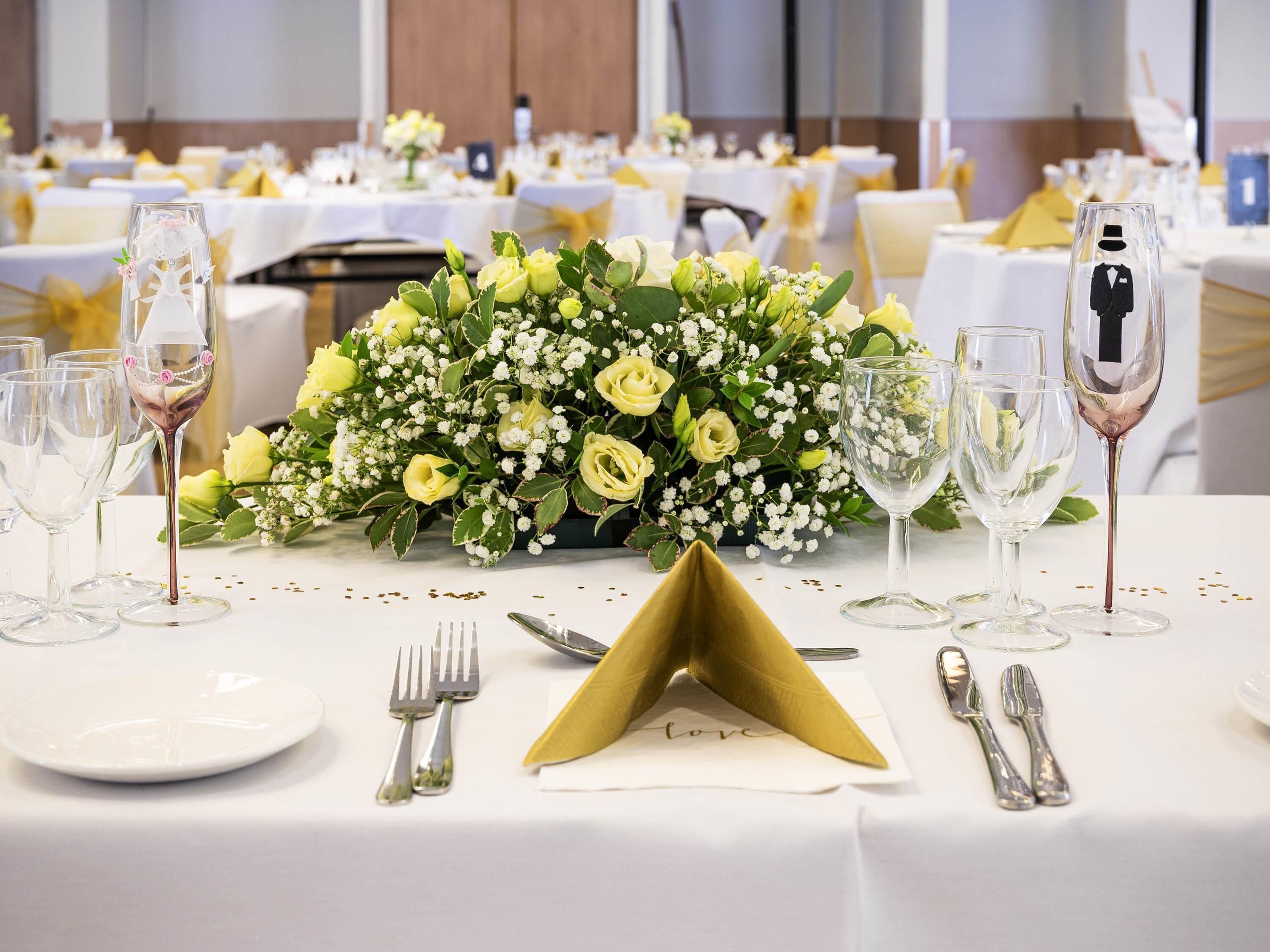 A banquet table set with white tablecloths, gold sashes, and a centerpiece of yellow roses and greenery.