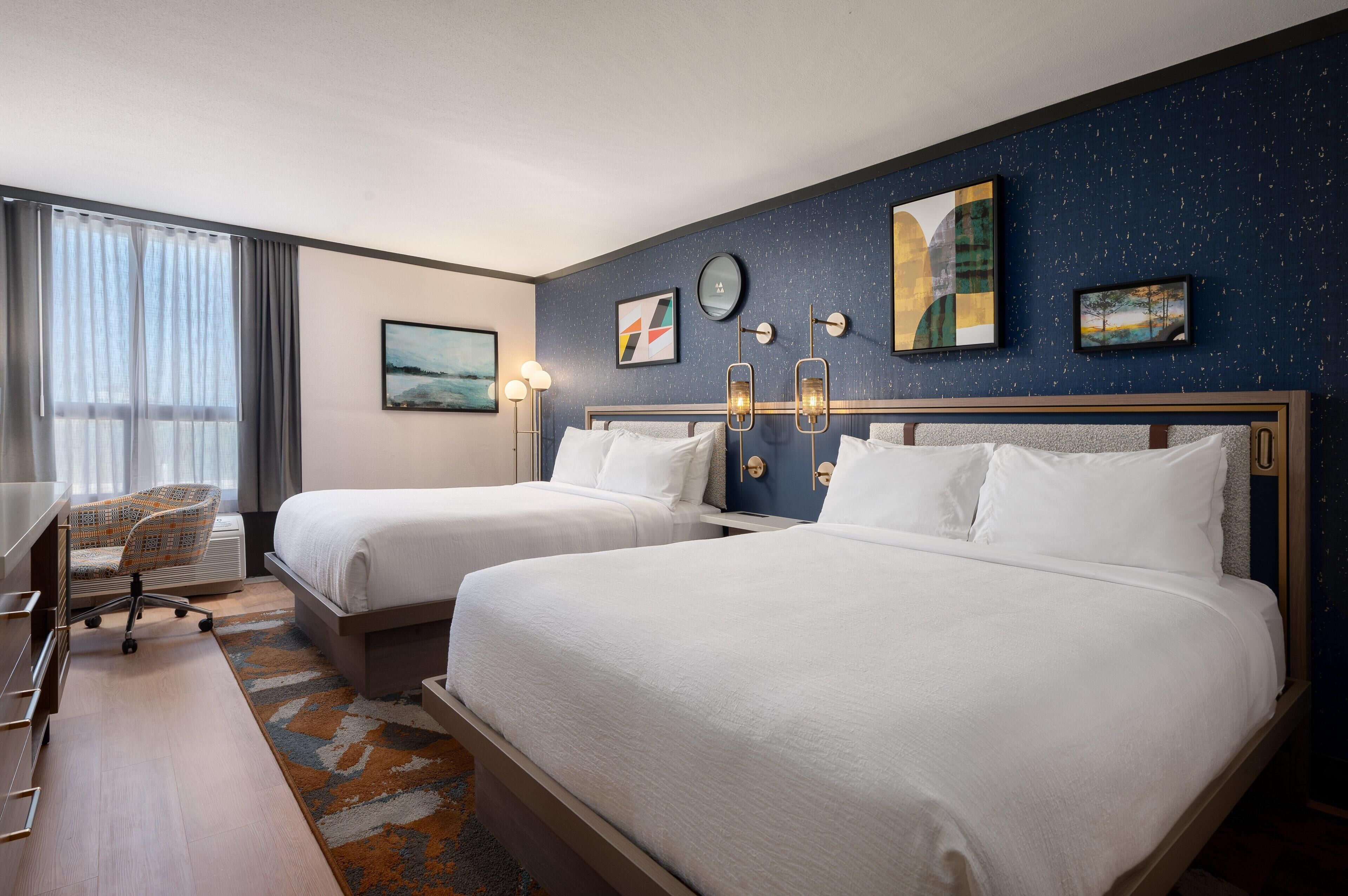 A hotel room with two beds, a desk, a chair, and artwork on the walls.