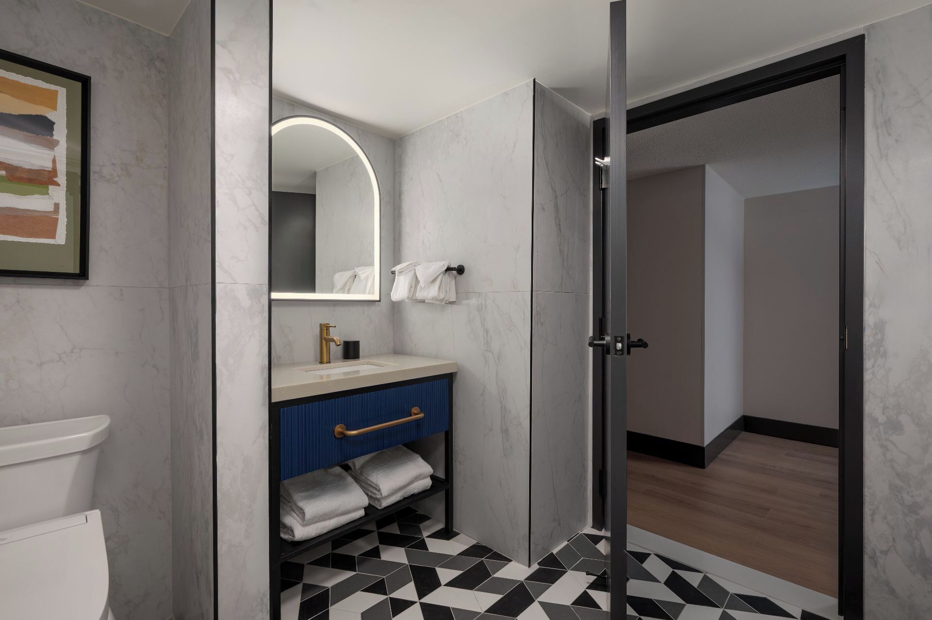 Un baño moderno con pared de mármol, lavamanos con grifo dorado, toallero azul y suelo de baldosas blanco y negro.