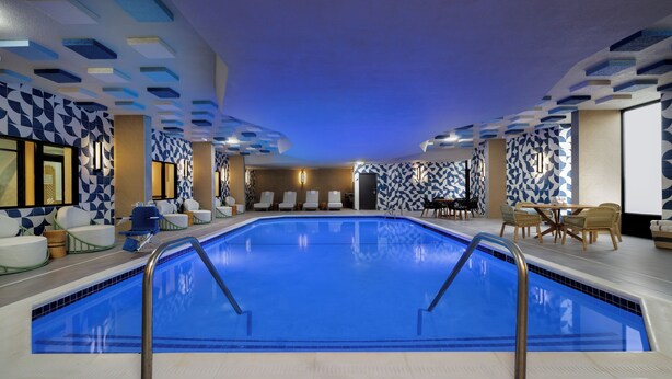 Une piscine intérieure avec un sol carrelé de bleu, entourée de chaises longues et d’un espace salon agrémenté d’un mur à motifs.