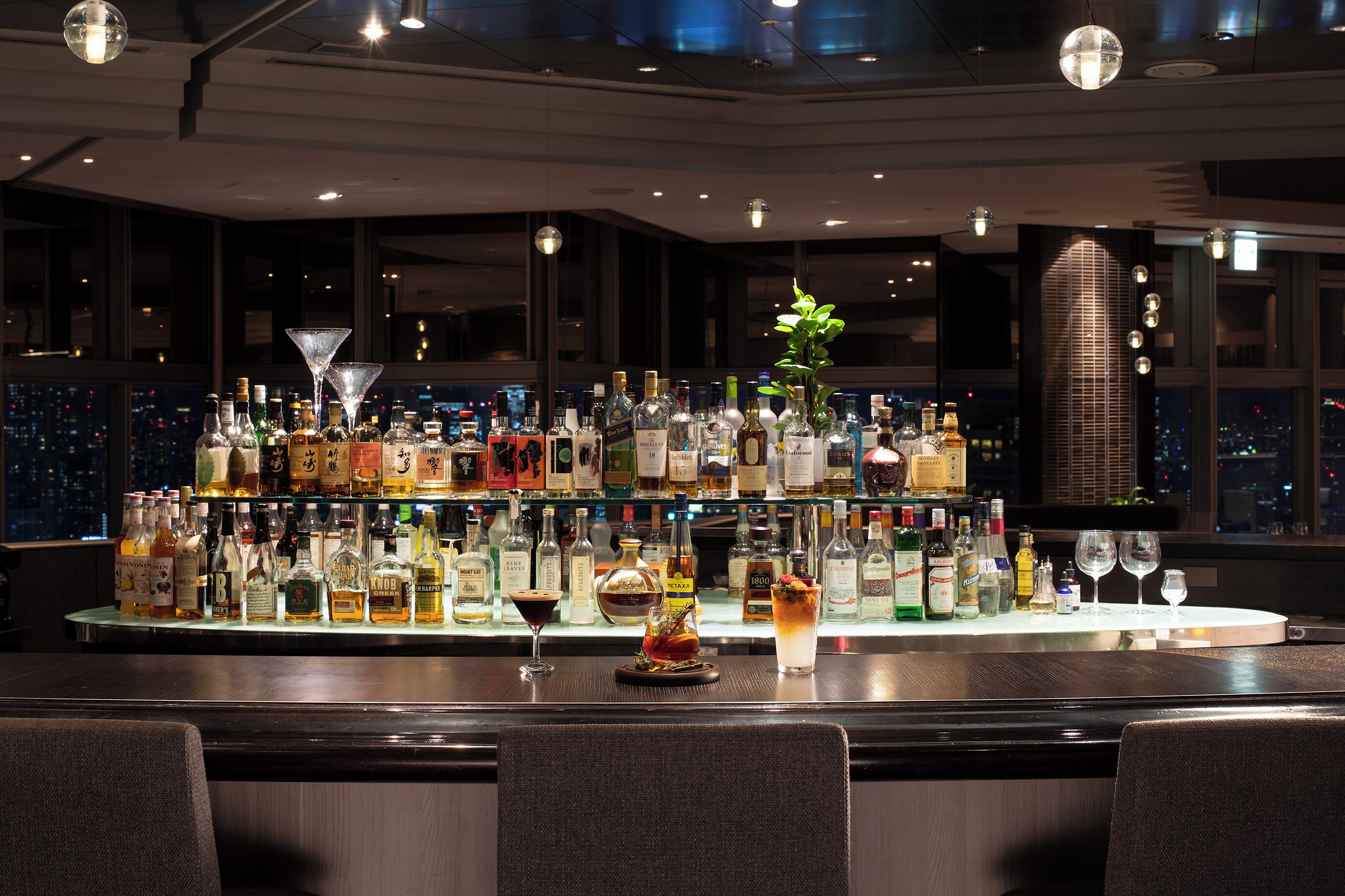 A bar area with a variety of liquor bottles and glasses, a decorative plant, and a city view through large windows.