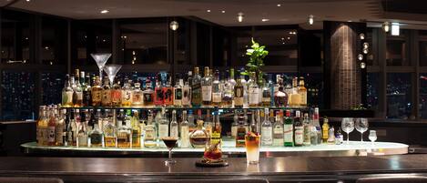 A bar area with a variety of liquor bottles and glasses, a decorative plant, and a city view through large windows.