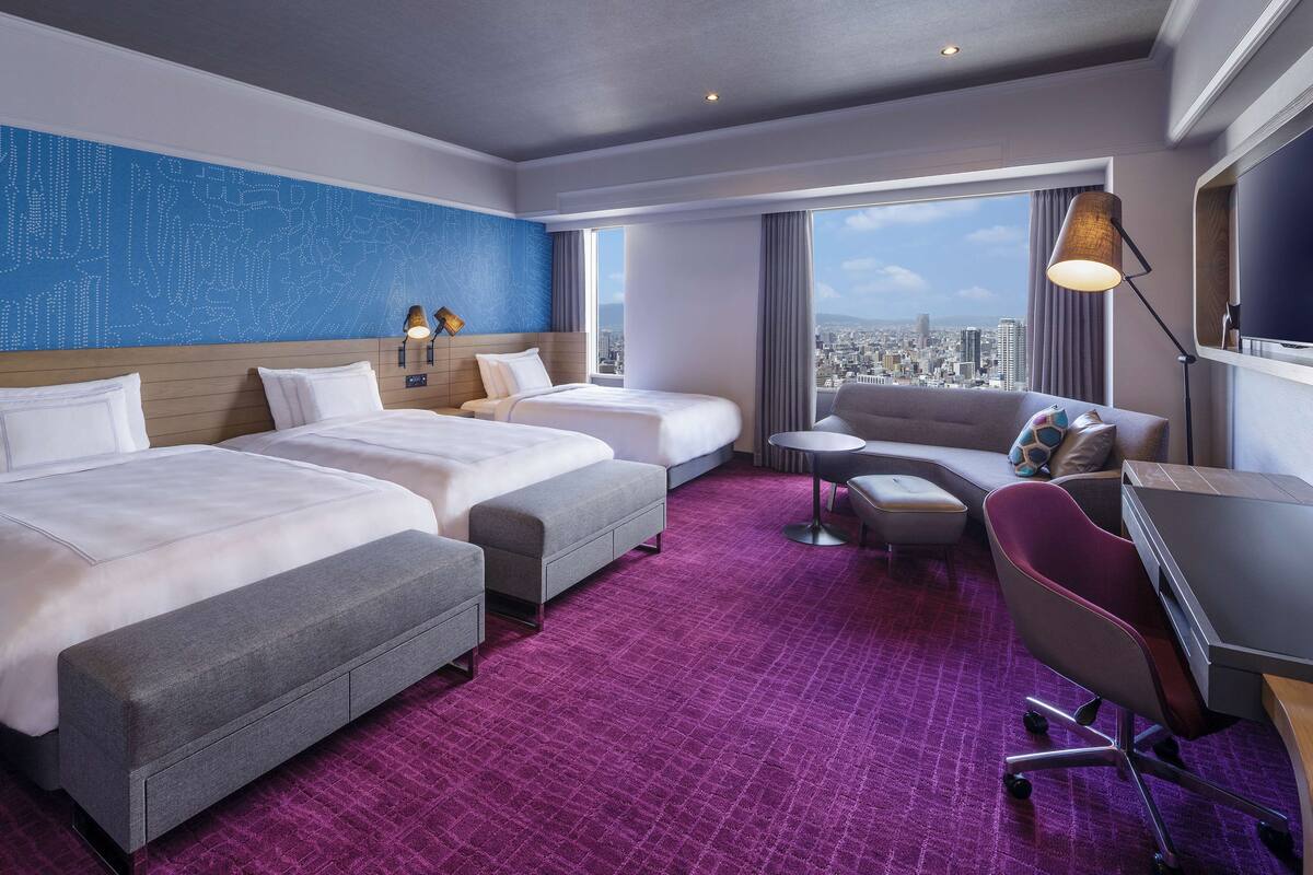 A hotel room with two beds, a sofa, a desk, and a large window offering a city view.