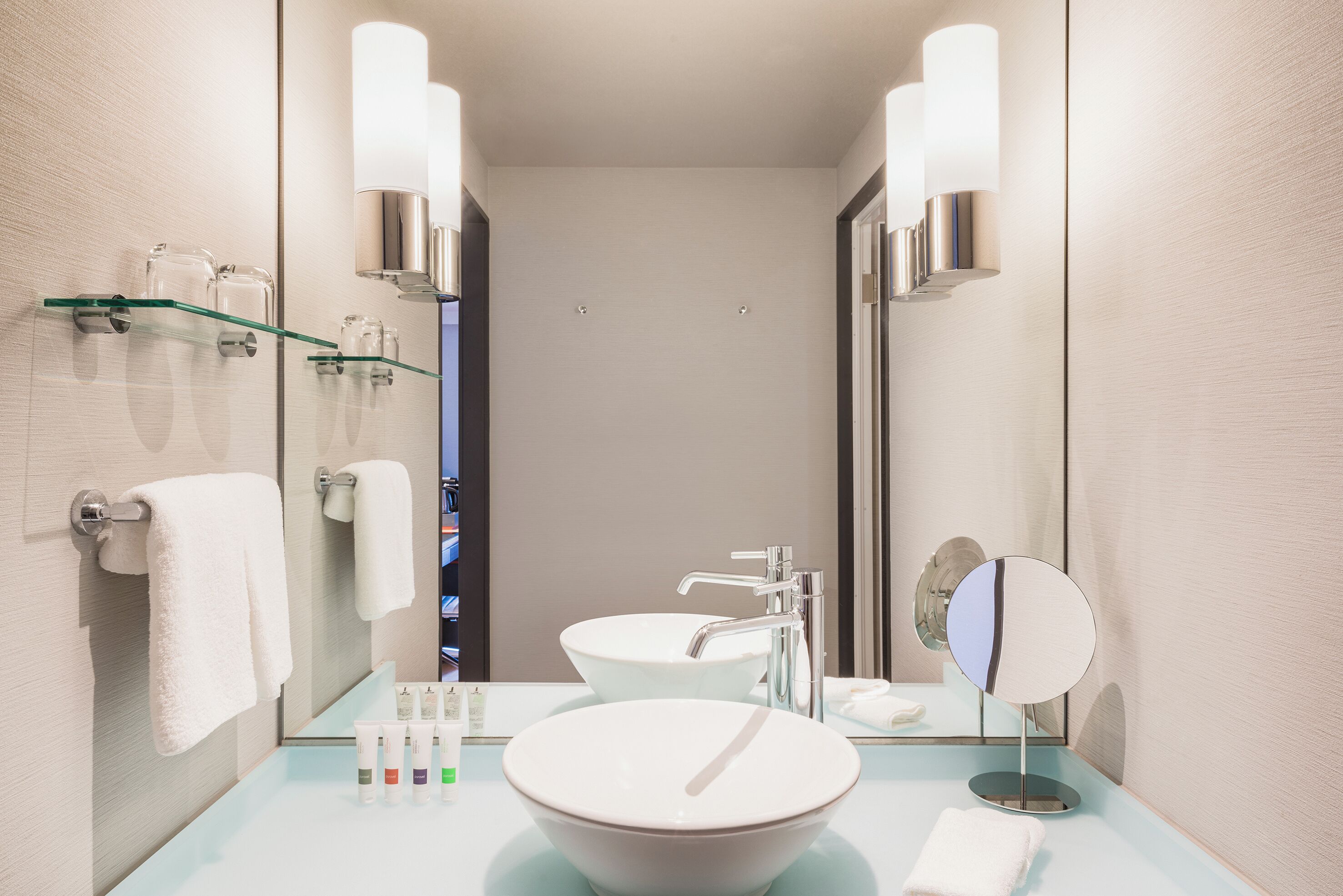 Un baño moderno con un espejo grande, un lavabo redondo y un estante de vidrio con artículos de aseo.