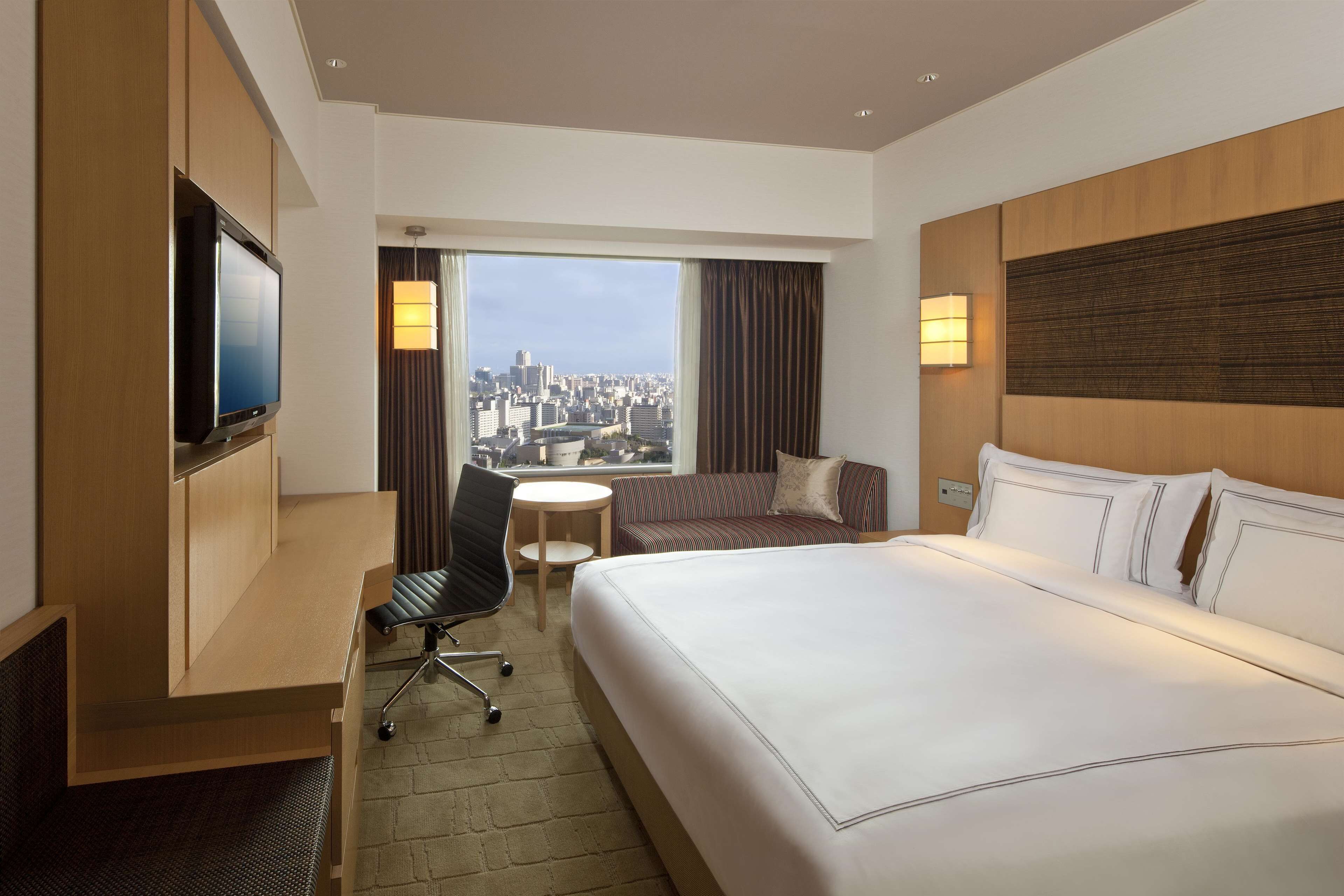 Hotelkamer met een groot bed, een bureau met stoel, een televisie, een bijzettafeltje en uitzicht op een stadsbeeld door grote ramen.