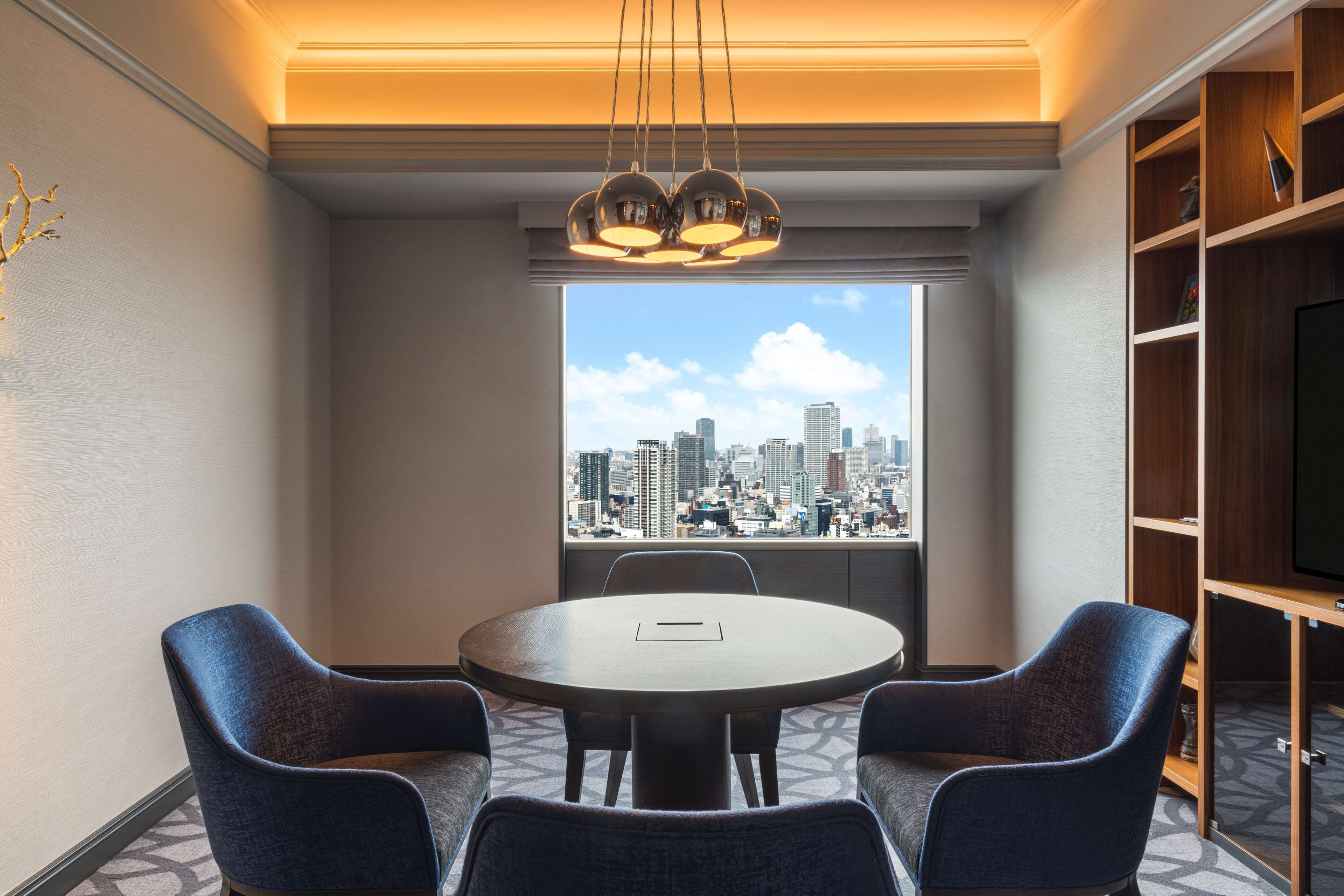 Habitación de hotel con vista a la ciudad, una mesa redonda con cuatro sillas, una estantería y un televisor.