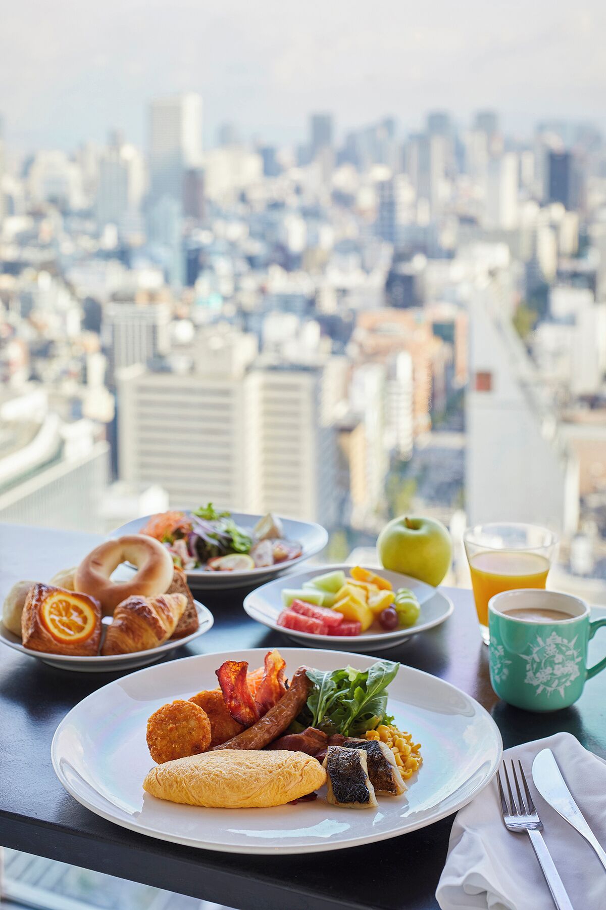 A breakfast setting with croissants, bacon, eggs, and fruit on a table with a city view in the background.