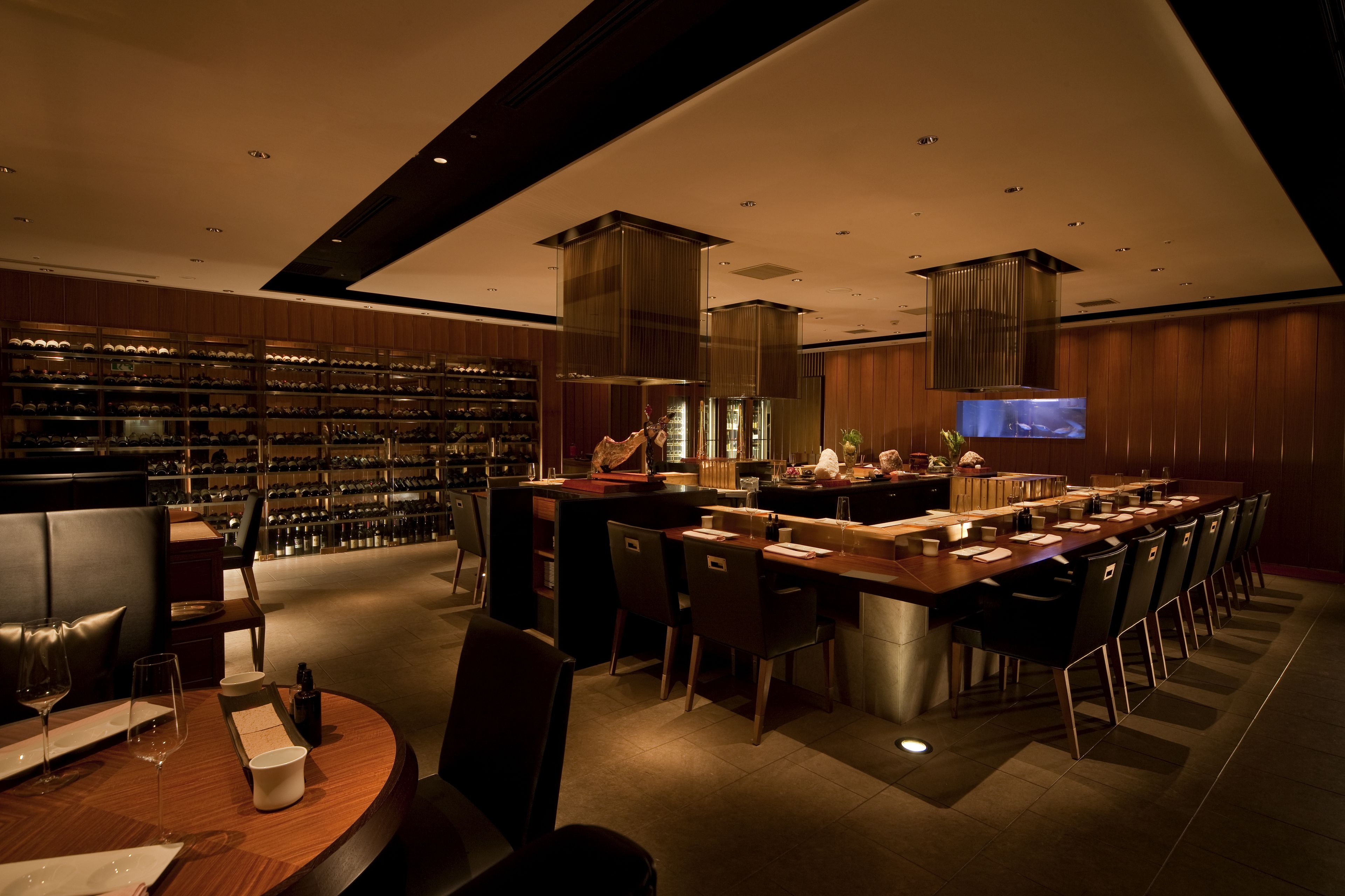A modern restaurant with a long dining table, multiple chairs, a bar area, and a wine rack.