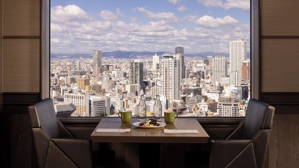 A dining area with a table set for two, a city view through large windows, and a plate of food. - Swissotel Nankai Osaka (Osaka)