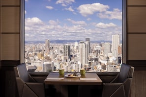 Un comedor con una mesa preparada para dos personas, vistas de la ciudad a través de amplias ventanas y un plato de comida.