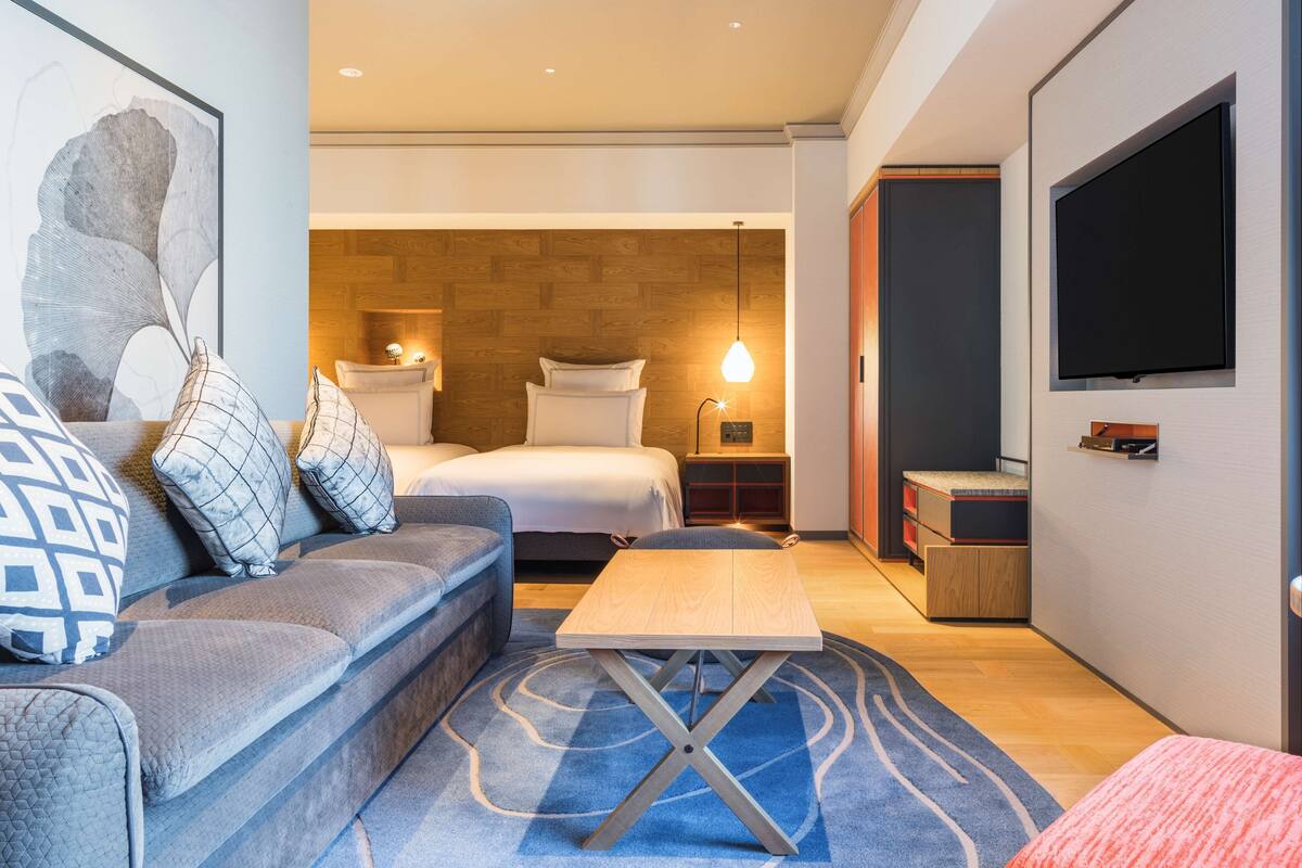 A modern hotel room with a bed, a sofa, a small table, and a TV mounted on the wall.