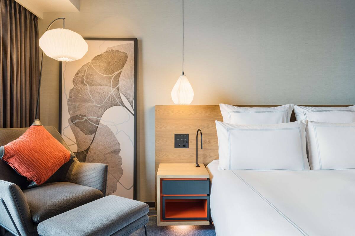A modern hotel room with a bed, a nightstand, a chair, and a wall art piece.
