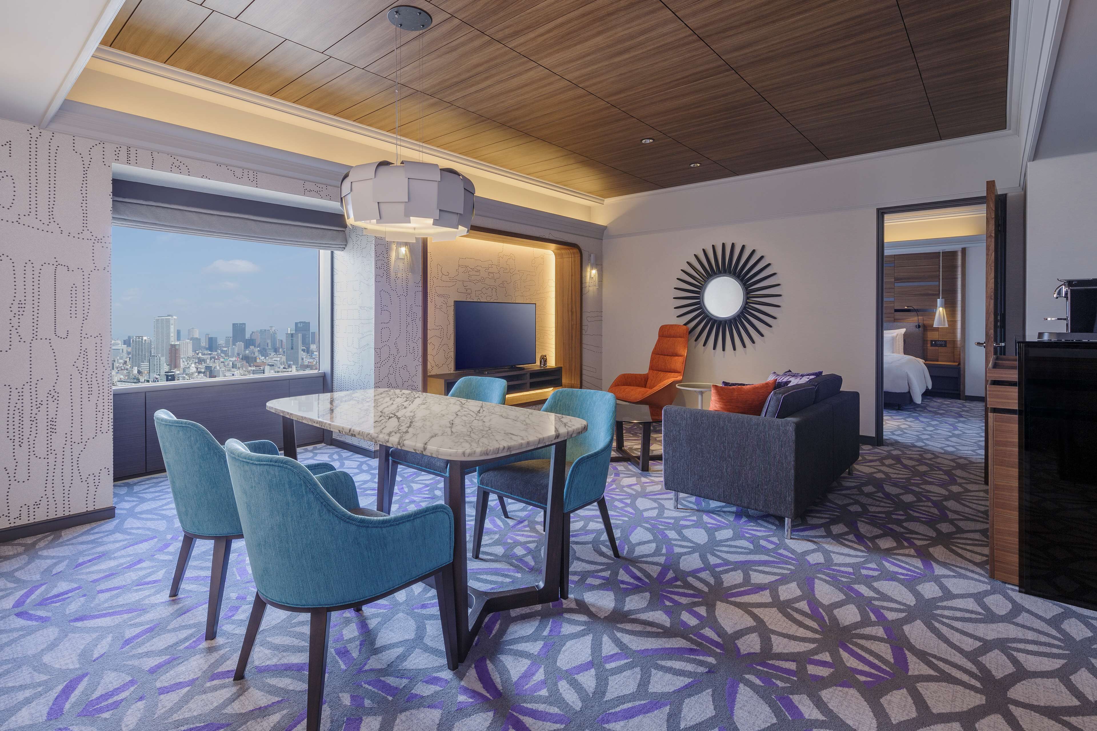 Ein modernes Hotelzimmer mit Blick auf die Stadt, ein Essbereich mit blauen Stühlen, eine Sitzecke und ein Fernseher.