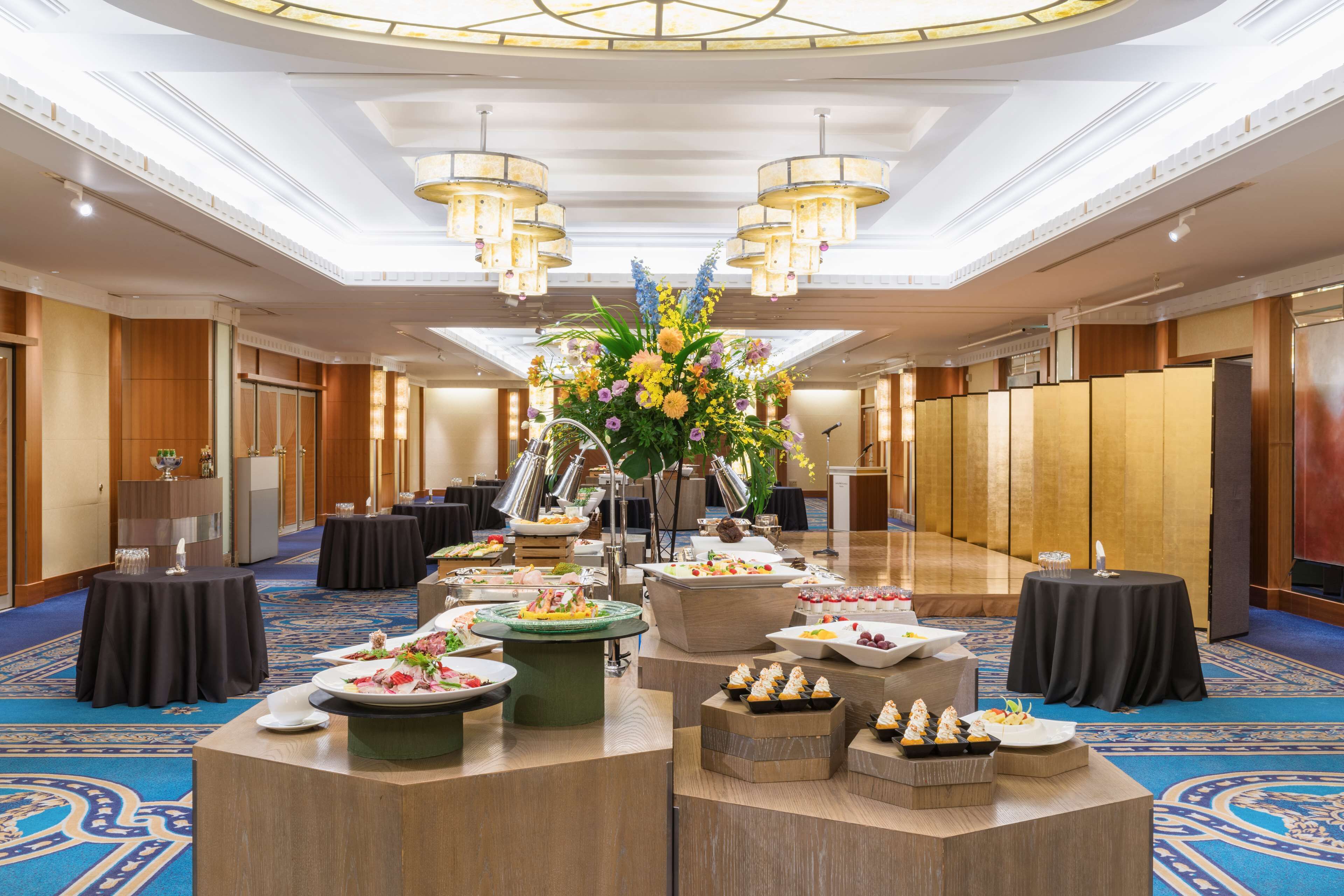 A spacious banquet hall with a central floral arrangement, multiple round tables, and a variety of food displays.