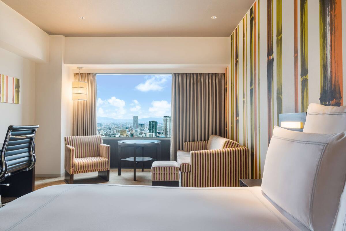 A hotel room with a large bed, two striped armchairs, a small table, and a view of a cityscape through large windows.