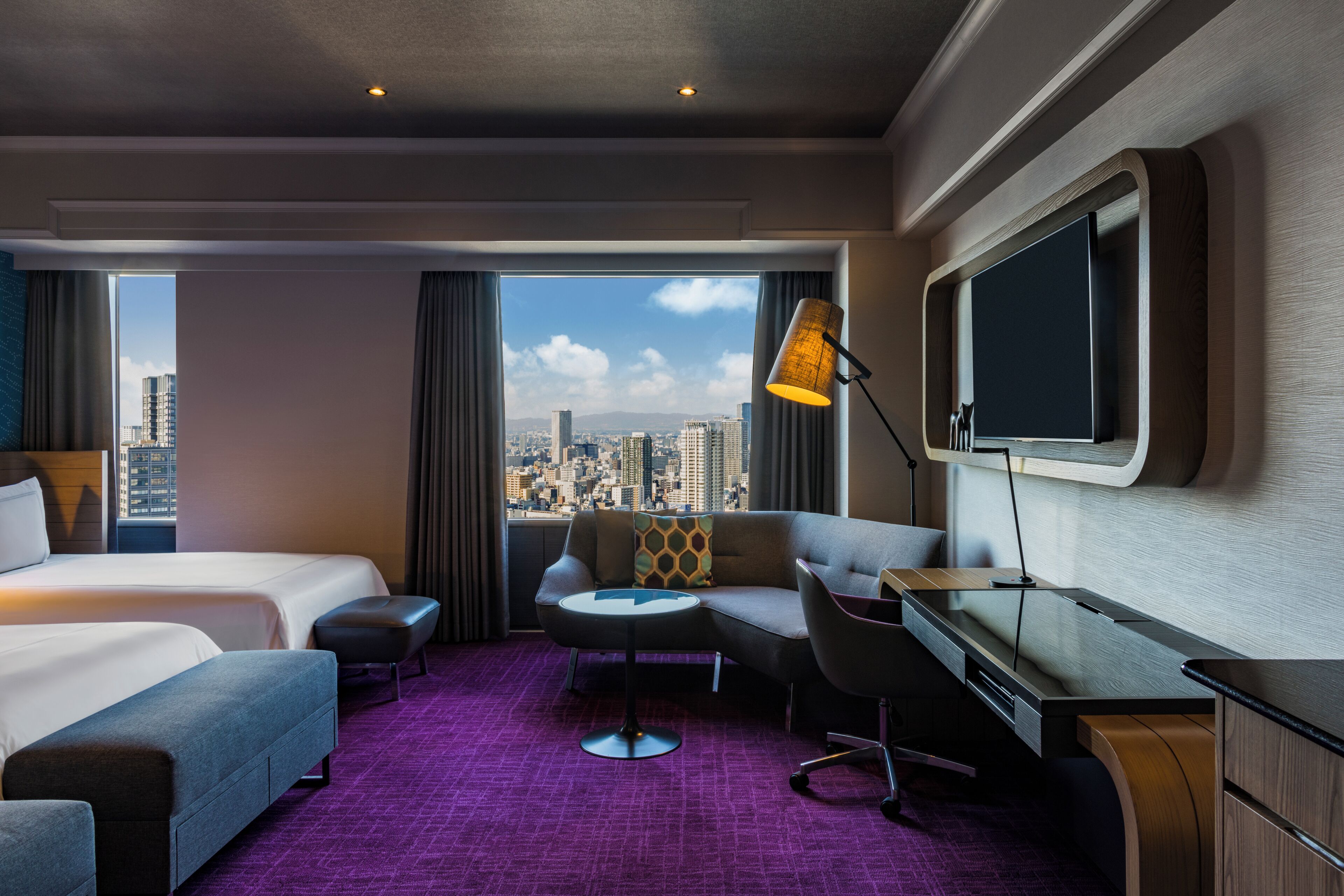 A hotel room with a bed, a desk, a chair, a small table, and a TV.