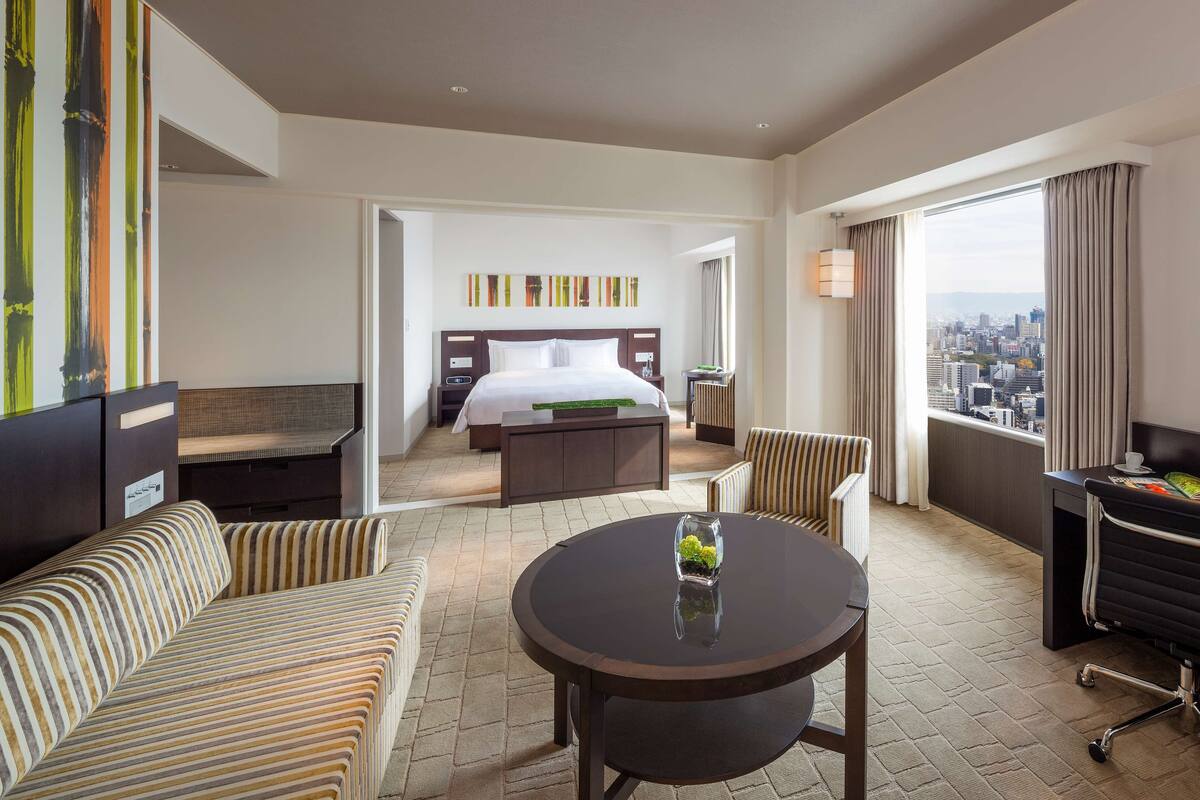 A hotel room with a bed, a sofa, a dining table, and a view of the city.