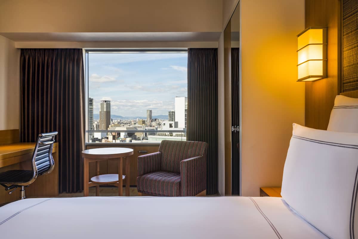 A hotel room with a large bed, a small table, a striped armchair, and a view of the cityscape through a large window.