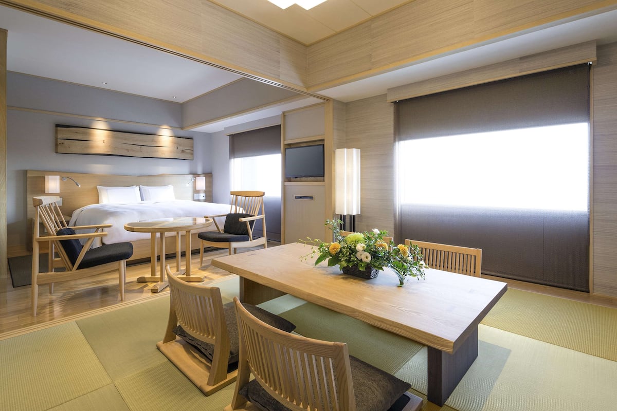 A modern hotel room with a bed, a dining table with chairs, a TV, and a large window.