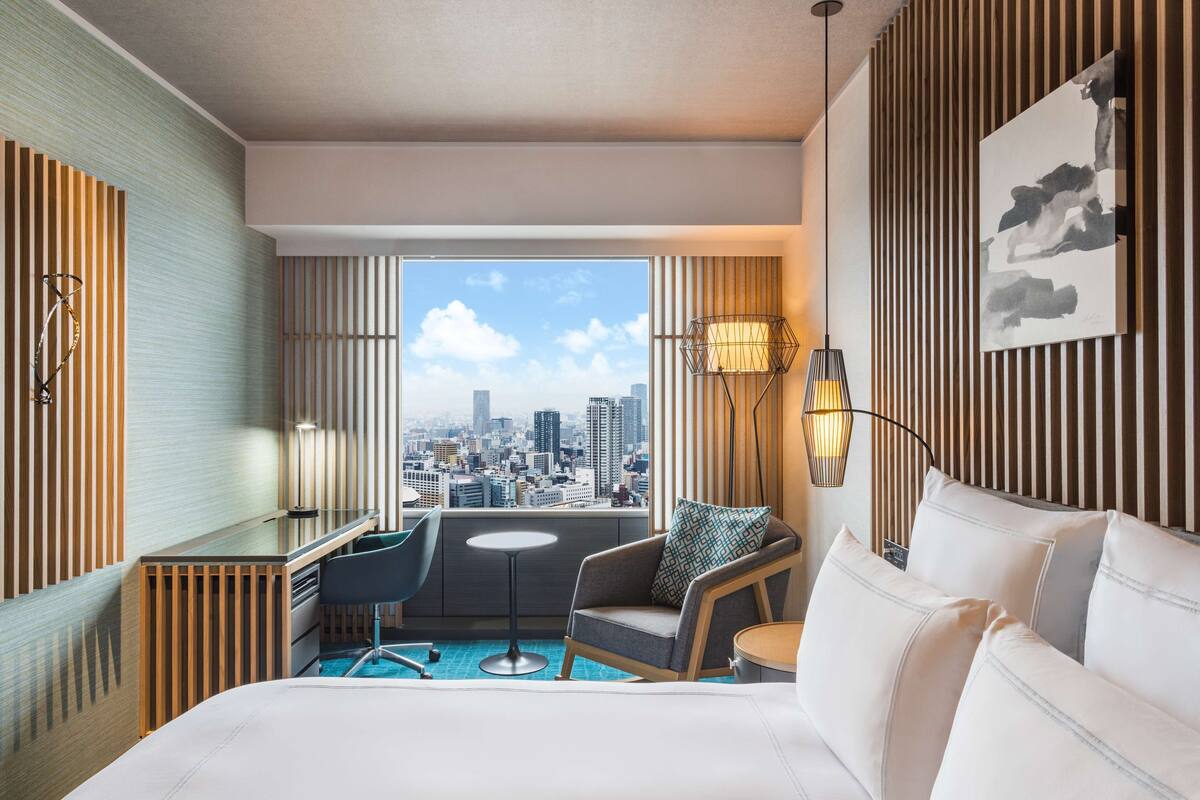 A modern hotel room with a city view, a desk, a chair, a bed, and a small table.