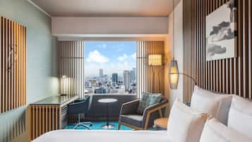 A modern hotel room with a city view, a desk, a chair, a bed, and a small table.