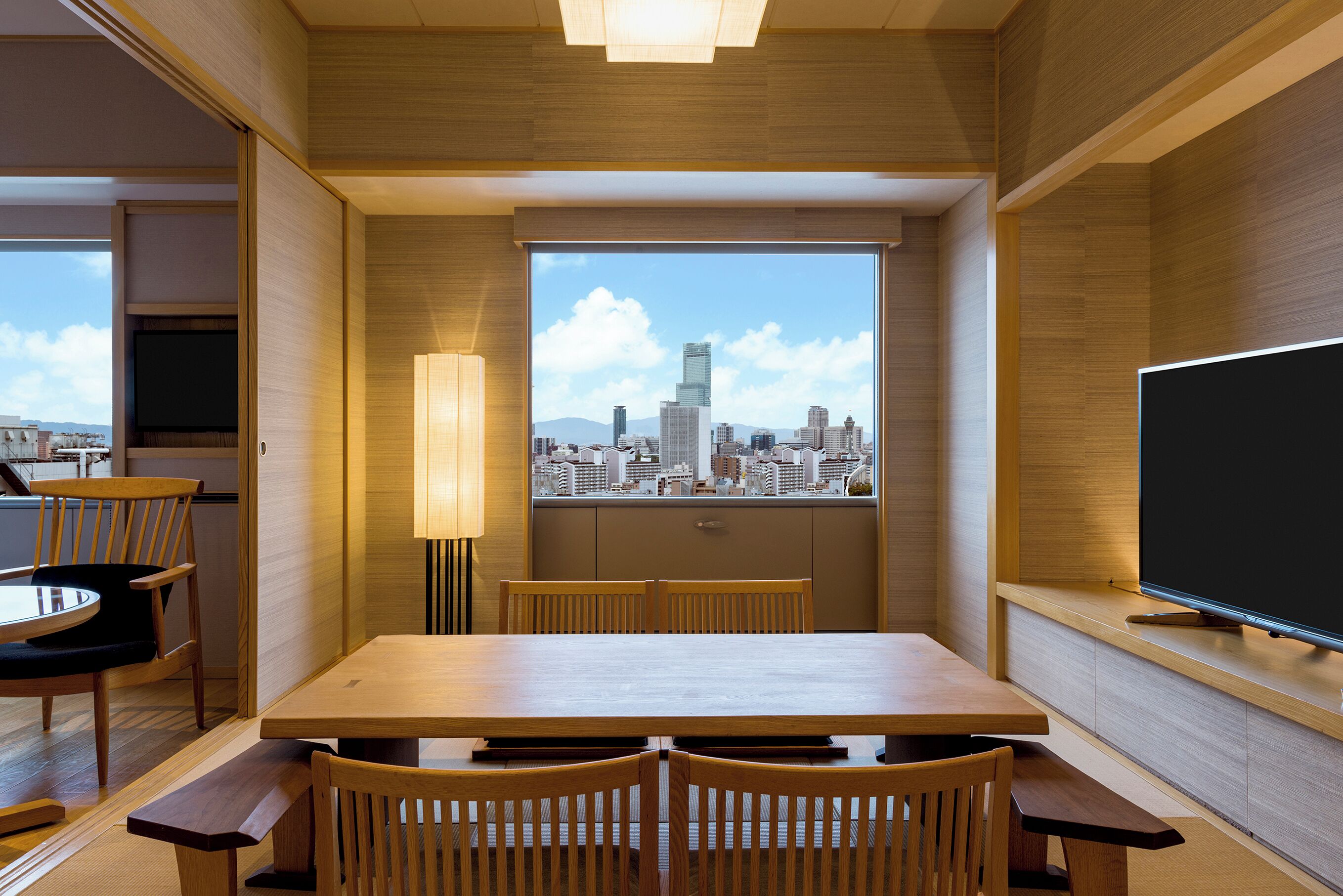 Una habitación moderna con una mesa y sillas de madera, un ventanal con vistas a la ciudad y un televisor de pantalla plana en la pared.