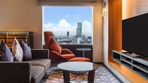 A modern hotel room with a sofa, armchair, and a small table. A large TV is mounted on the wall, and there's a window offering a city view. - Swissotel Nankai Osaka (Osaka)