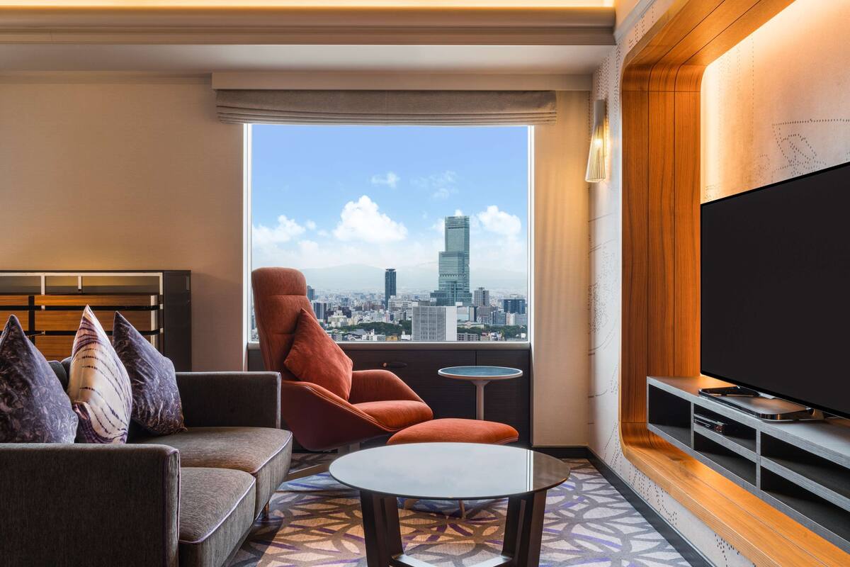 A modern hotel room with a sofa, armchair, and a small table. A large TV is mounted on the wall, and there's a window offering a city view.