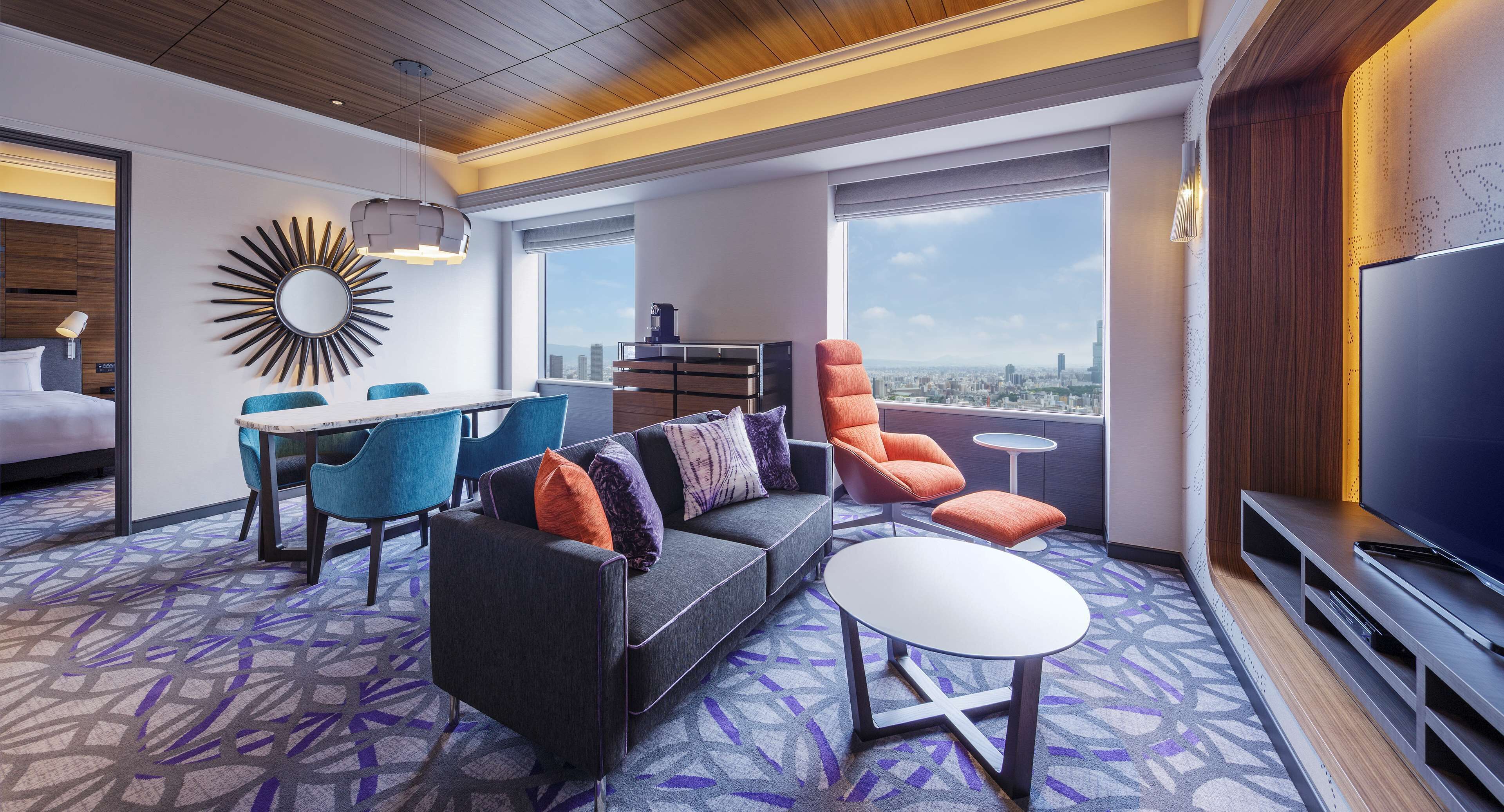 Una habitación de hotel moderna con sofá, sillas, una mesa de comedor, televisor y vistas a la ciudad.