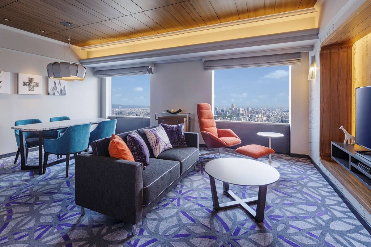 A modern hotel room with a city view, a dining area, and a seating area with a sofa and chairs.