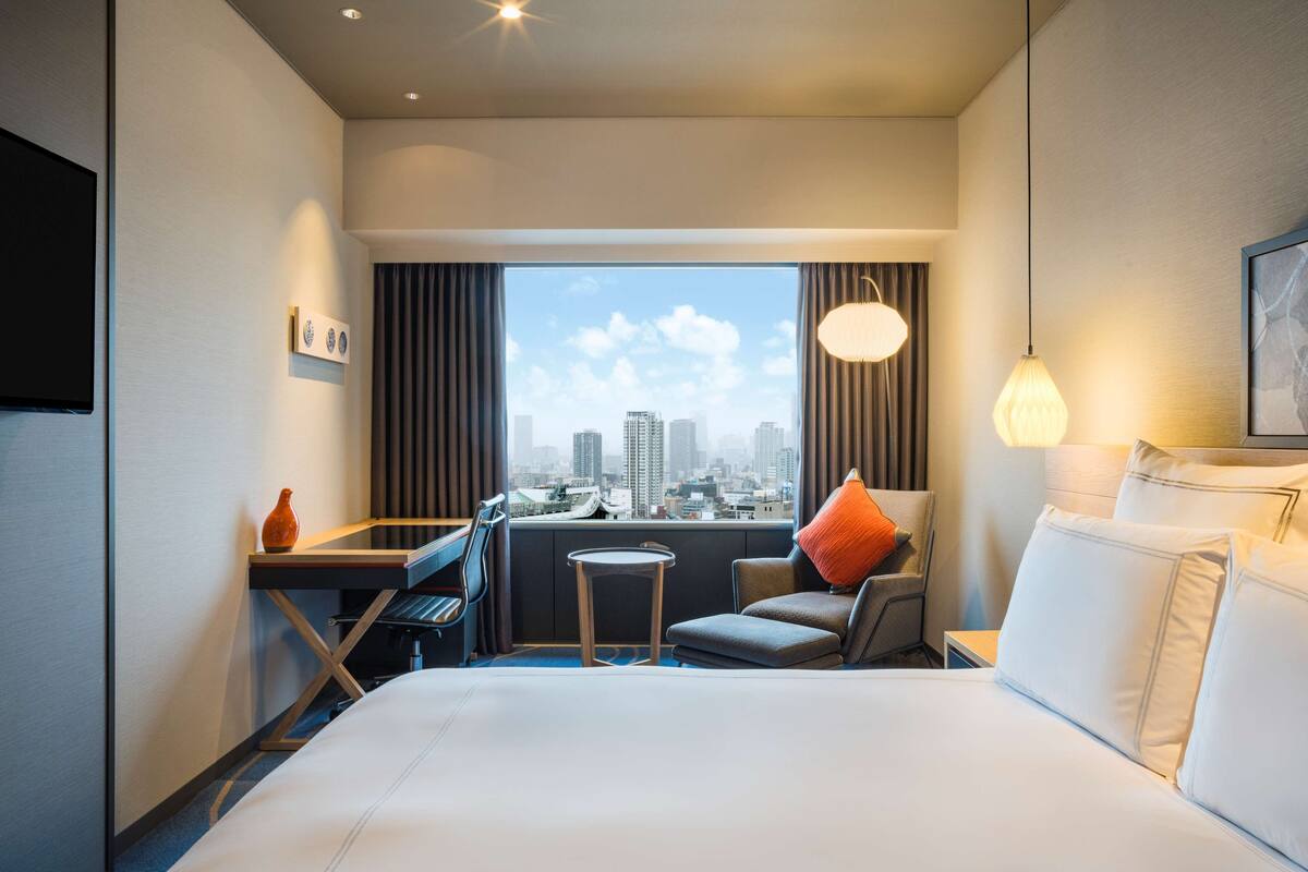 A hotel room with a bed, a desk, a chair, a small table, and a view of the city skyline through large windows.