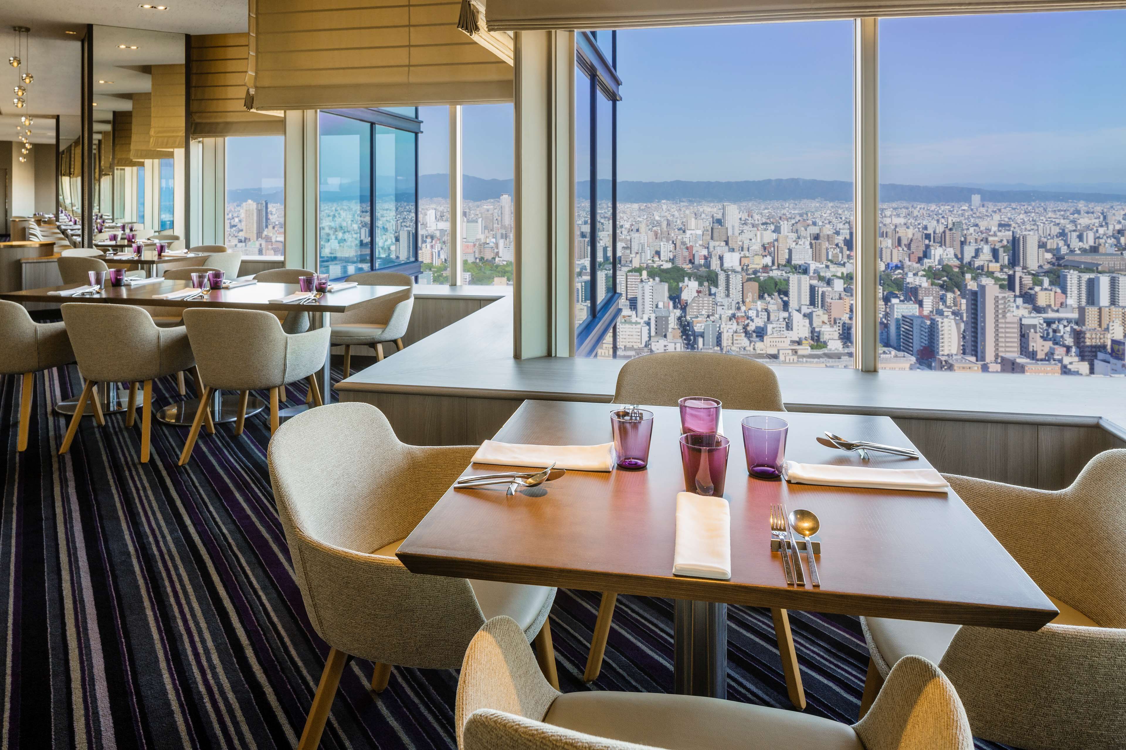 Un comedor moderno con mesas y sillas, ventanas amplias con vista a la ciudad y una alfombra a rayas.