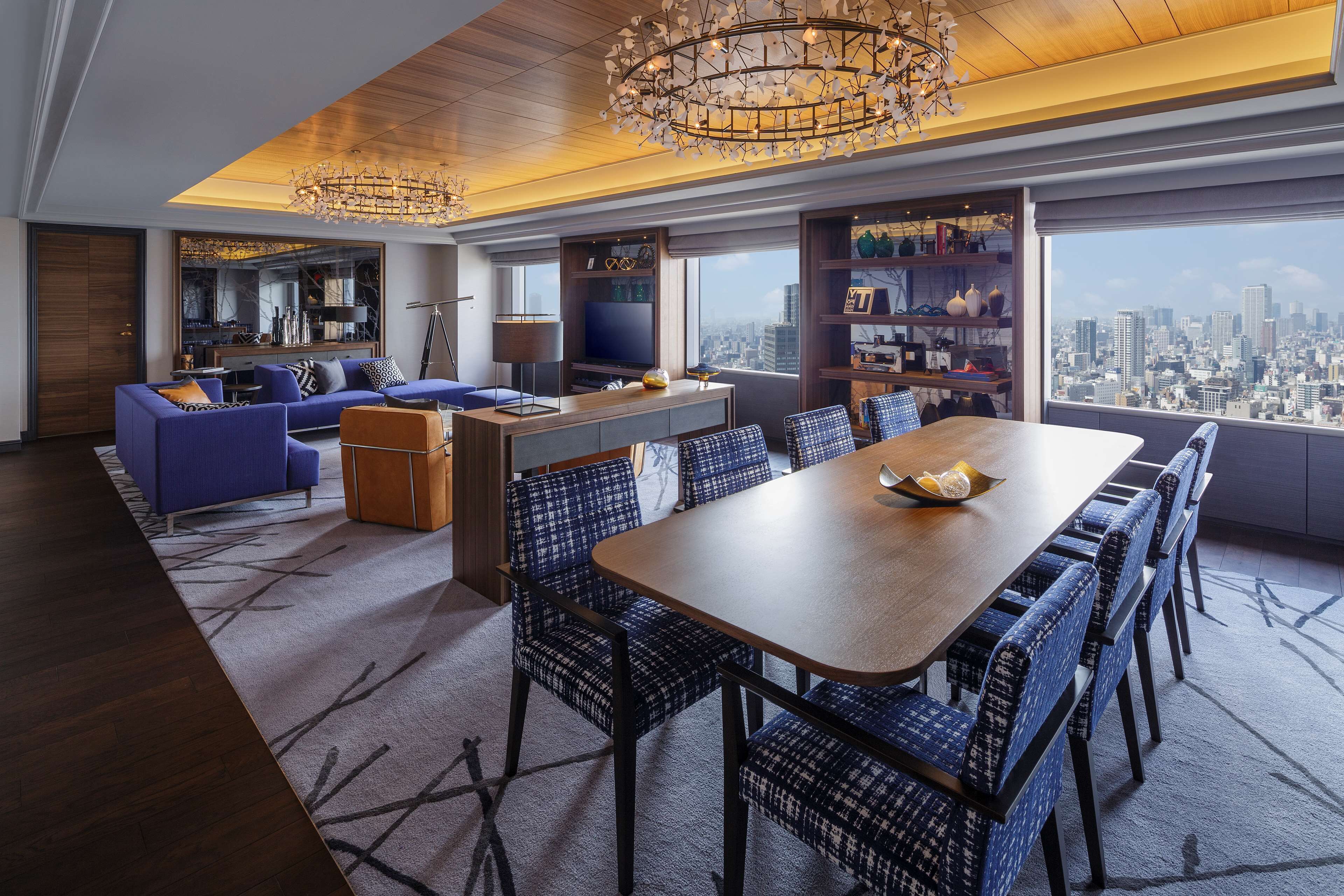 A modern living room with a large dining table, blue upholstered furniture, and a city view through large windows.