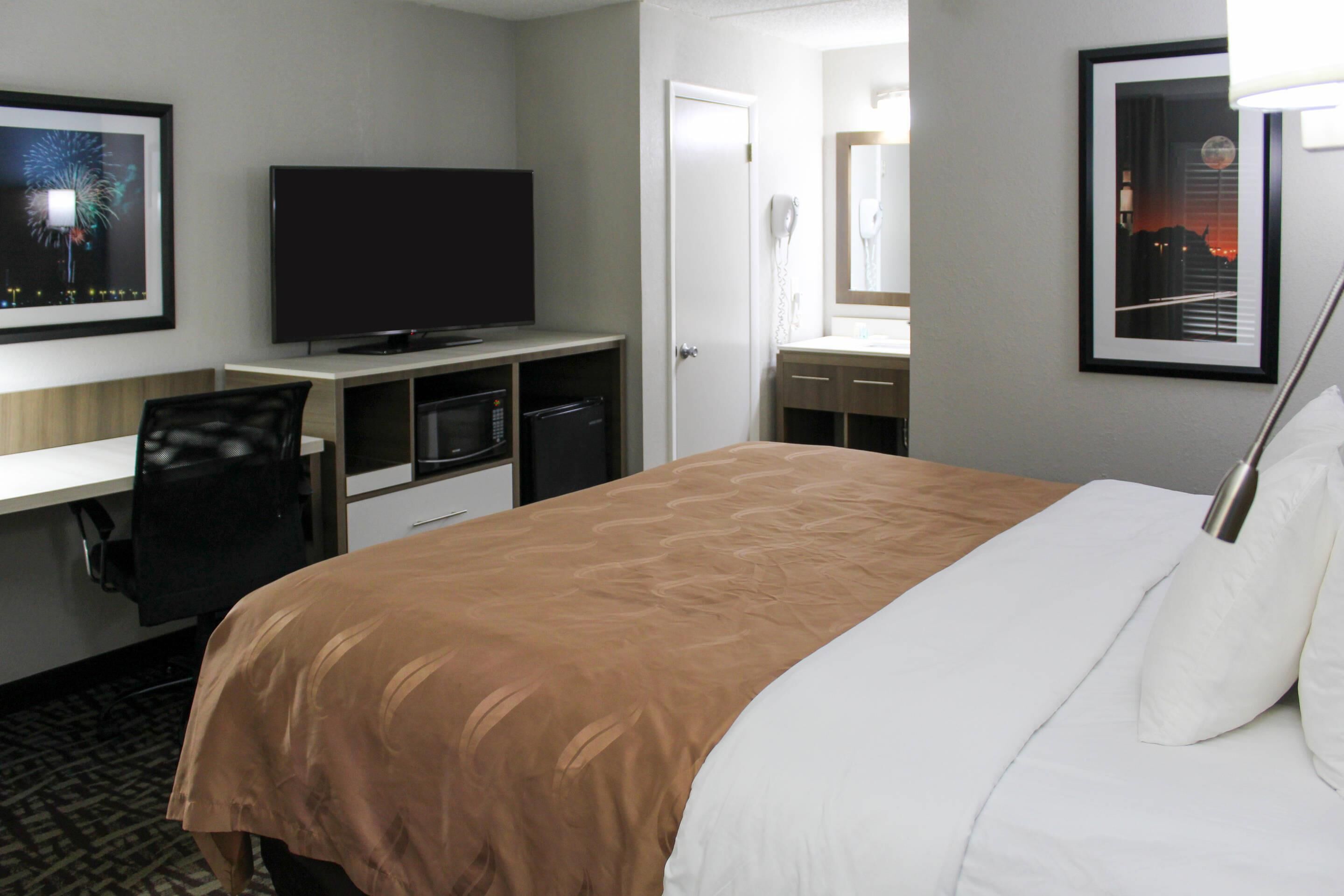 A hotel room with a bed, a TV, a desk, a chair, a mirror, and a framed picture on the wall.