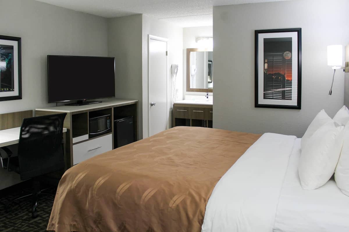 A hotel room with a bed, a desk with a flat-screen TV, a mirror, and a framed picture on the wall.